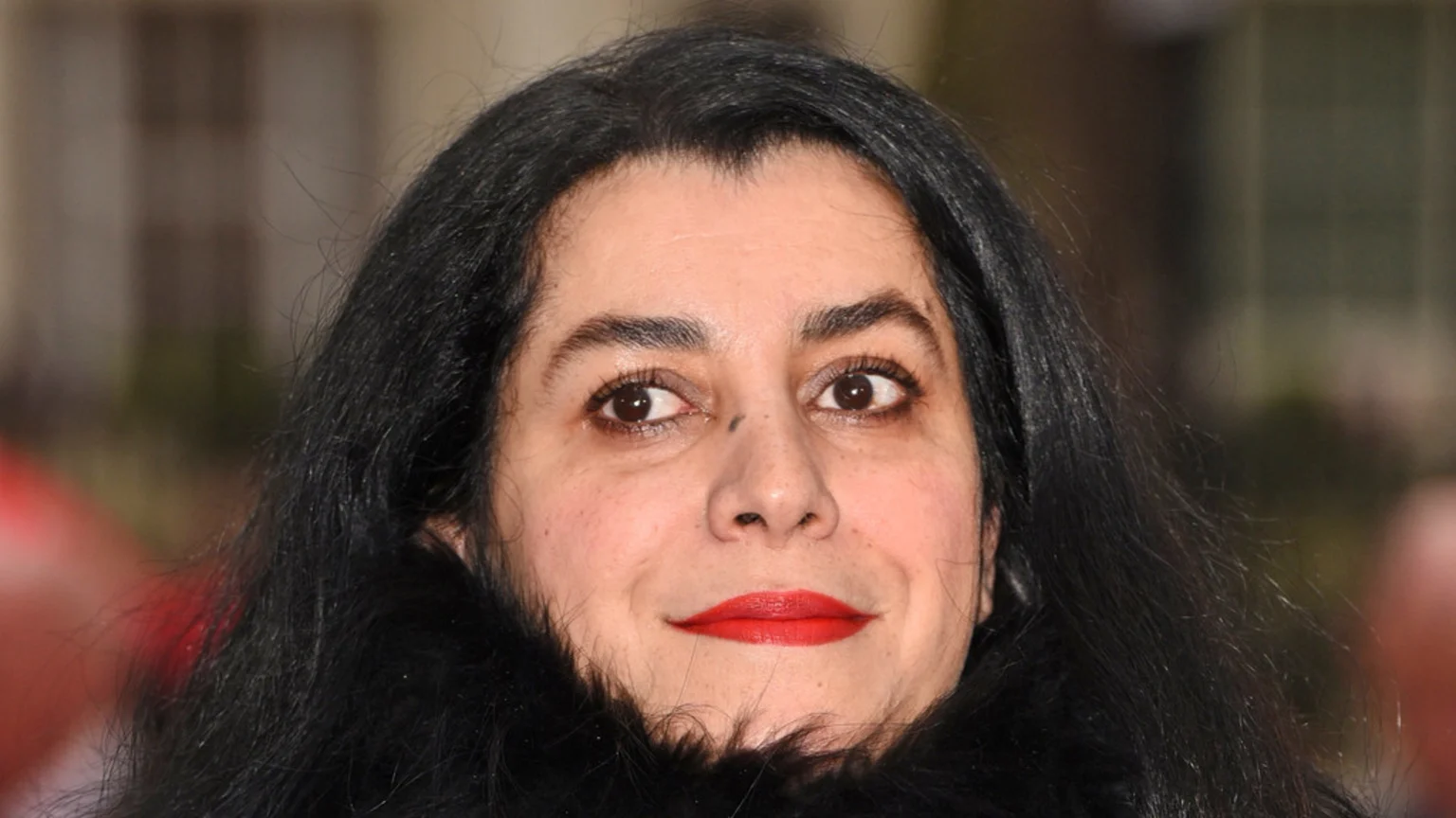A close-up of a woman with long dark hair, dark eyes, and red lipstick, looking towards the viewer with a slight smile.