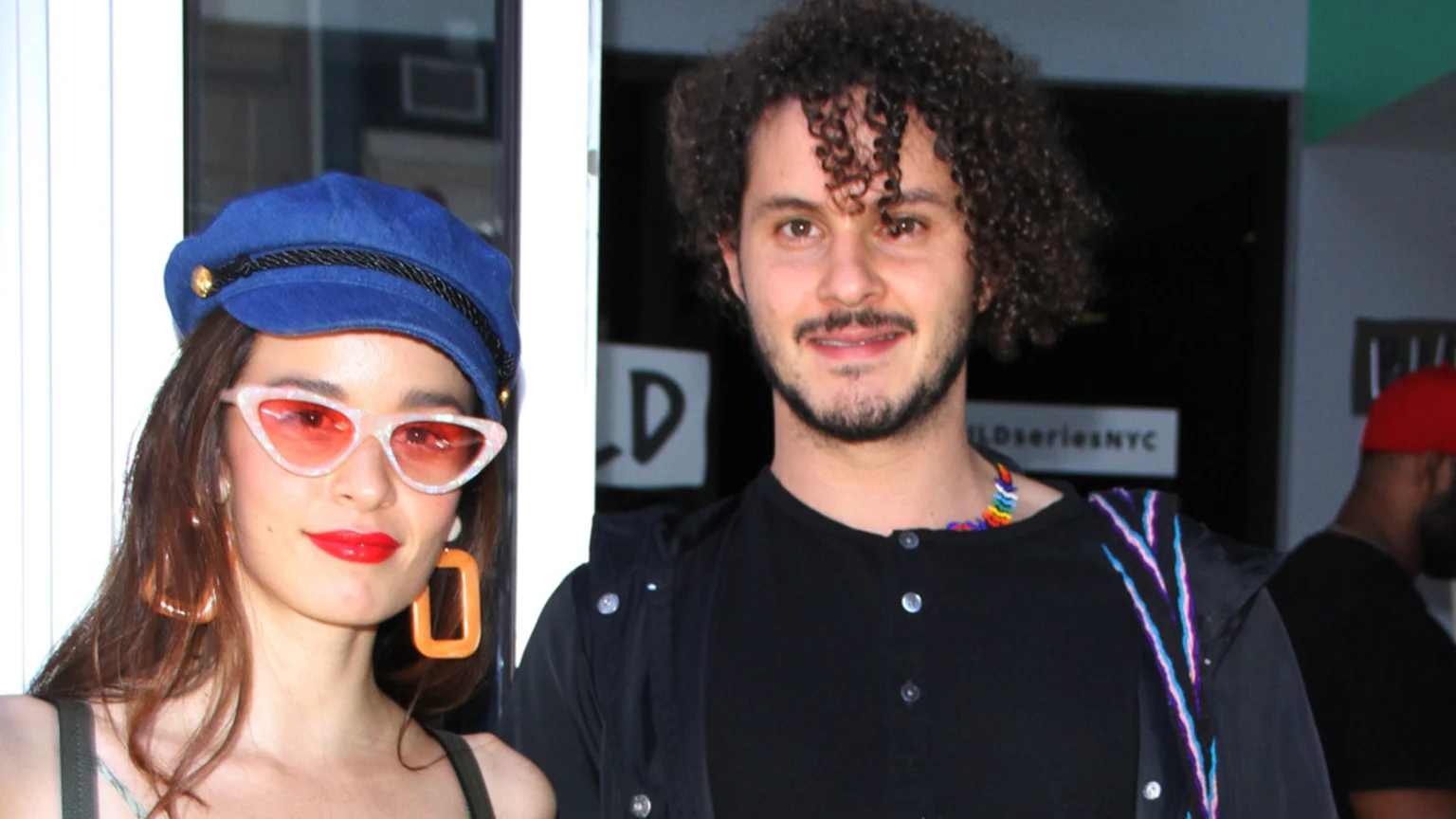 A woman in a blue captain's hat, pink cat-eye sunglasses, and red lipstick smiles next to a man with curly hair and a beard.