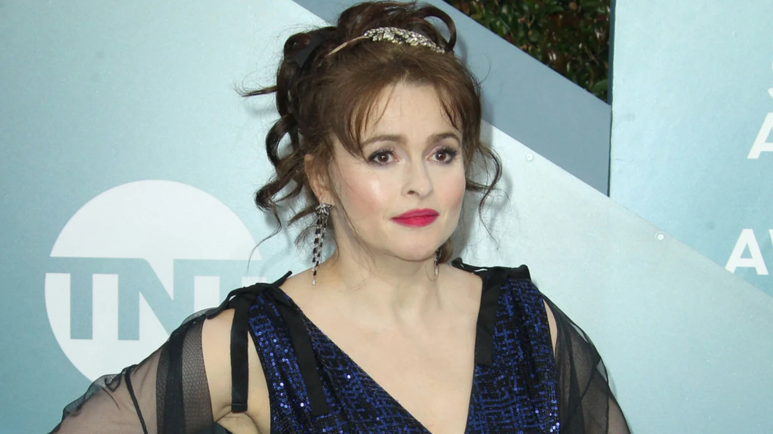 Helena Bonham Carter poses at an event, wearing a dark blue sequined dress, long earrings, and a sparkly headband, with her hair styled up.