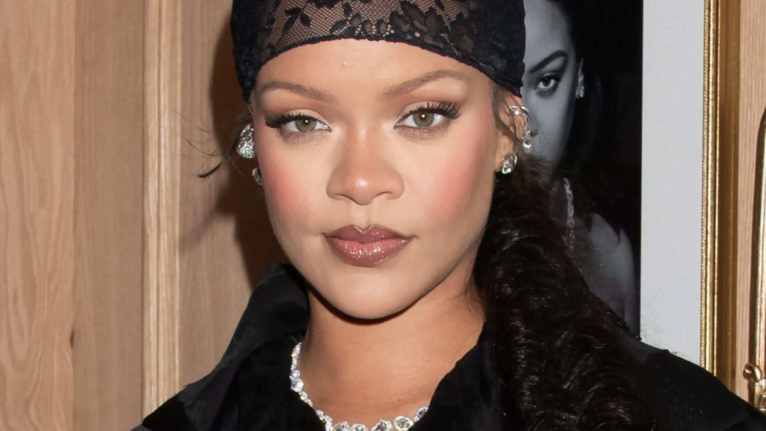 Close-up of Rihanna looking directly at the camera, wearing a black lace headscarf, prominent diamond earrings and necklace. A framed black and white photo is partially visible behind her right side.