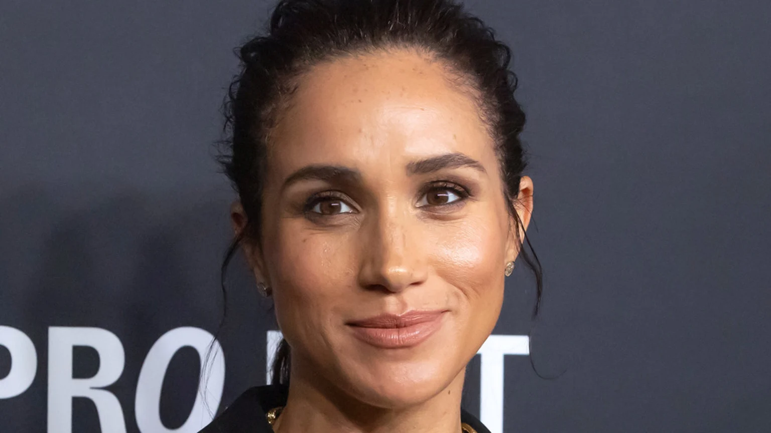 Close-up of Meghan, Duchess of Sussex, smiling slightly, with her dark hair pulled back and wearing light makeup, against a dark background with white text visible.