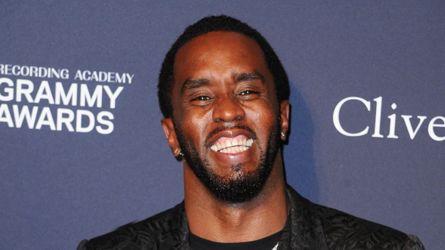 Sean 'Diddy' Combs, a man with a beard and short dark hair, smiles broadly at the camera, wearing a dark patterned jacket, with "Grammy Awards" and "Clive" text partially visible in the dark background.