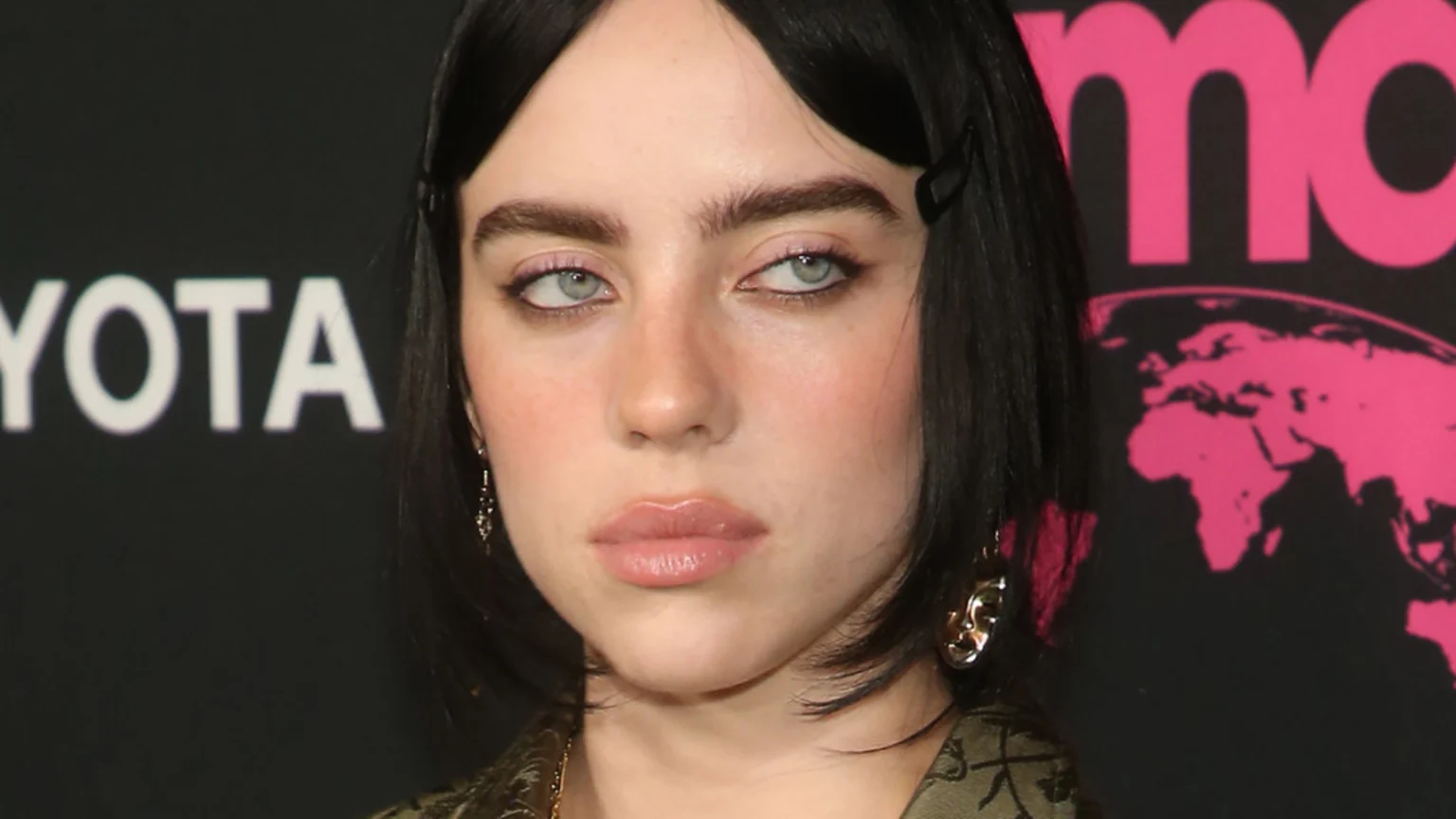 Close-up of Billie Eilish with dark hair and light eyes, wearing pink eye makeup, looking slightly to the side against a dark background with partial white and pink logos.