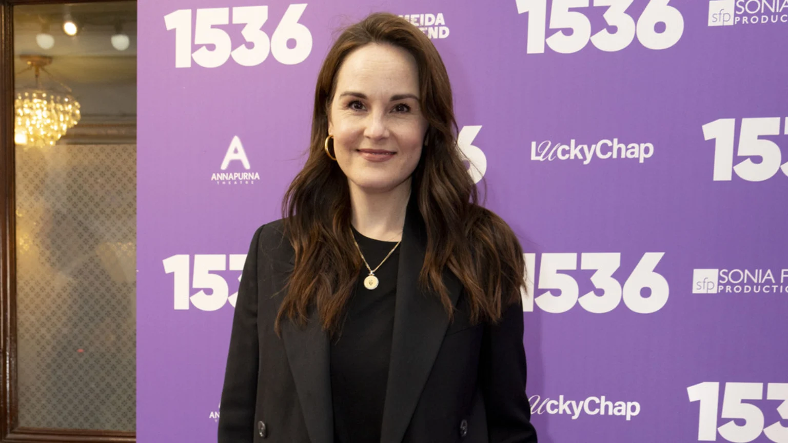 Michelle Dockery smiles at the camera, wearing a black blazer and a gold necklace, standing in front of a purple backdrop with "1536" and various production logos.