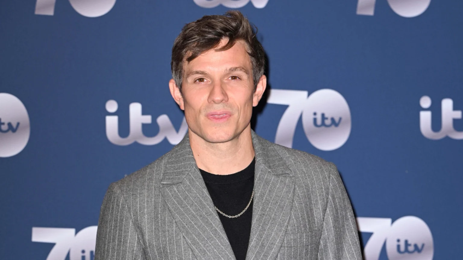 Will Best stands in front of a blue background with white ITV 70th anniversary logos, wearing a grey pinstripe blazer, a black top, and a silver chain, looking at the camera.