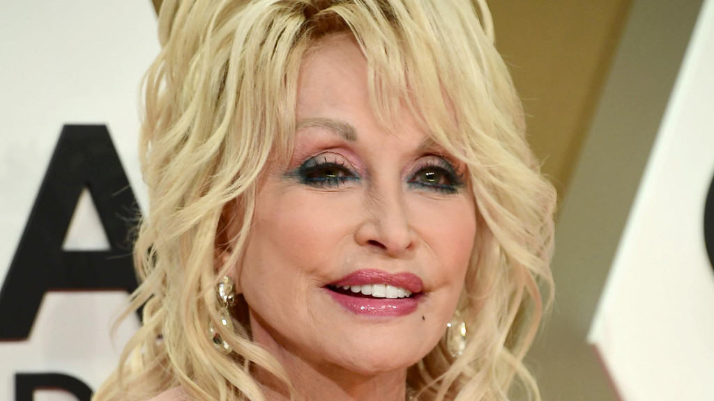 Dolly Parton smiles in a close-up, featuring her blonde hair, prominent pink and blue eye makeup, and glossy pink lips.