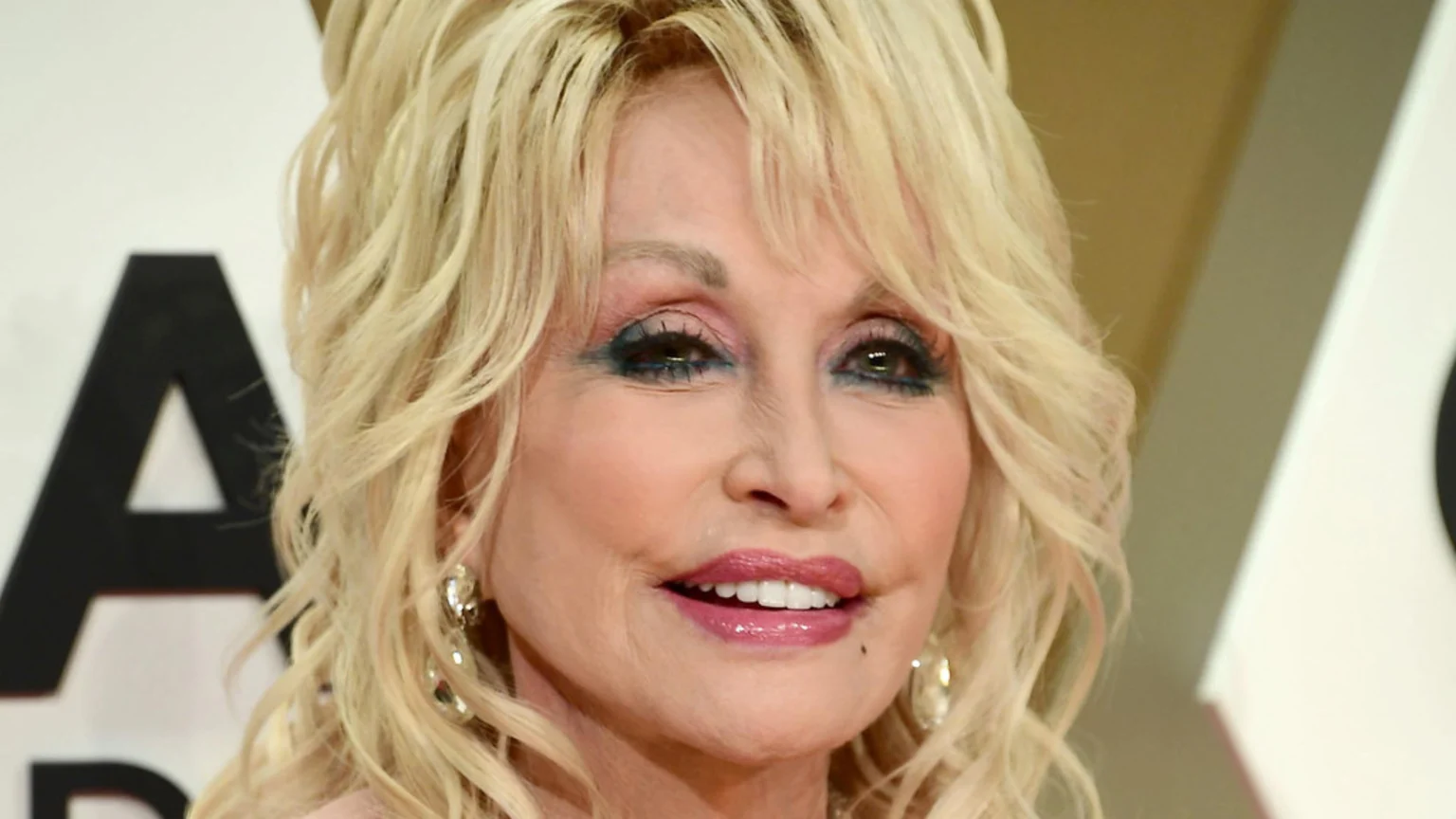 Dolly Parton smiles in a close-up, featuring her blonde hair, prominent pink and blue eye makeup, and glossy pink lips.