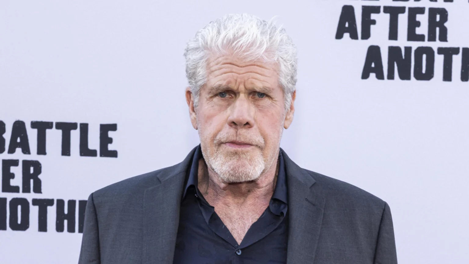 Actor Ron Perlman, with white hair and a goatee, looks directly at the camera, wearing a dark shirt and suit jacket, against a light background with partial black text.