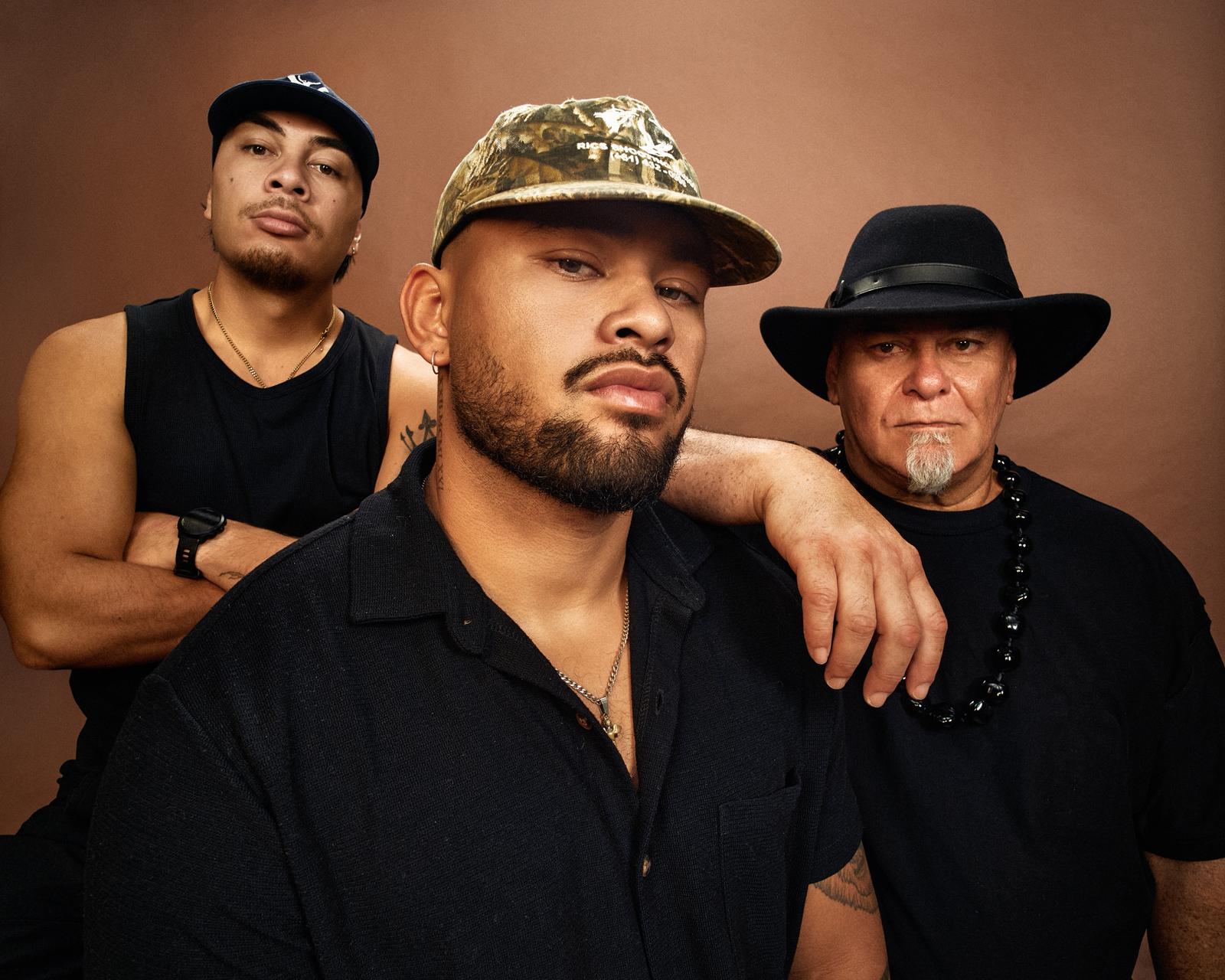 Three men in black pose seriously against a brown background; the left man wears a navy cap, the central man a bearded face and camo cap with his arm on the elder, who wears a black hat and beaded necklace.