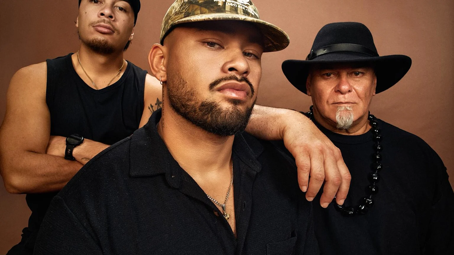 Three men in black pose seriously against a brown background; the left man wears a navy cap, the central man a bearded face and camo cap with his arm on the elder, who wears a black hat and beaded necklace.