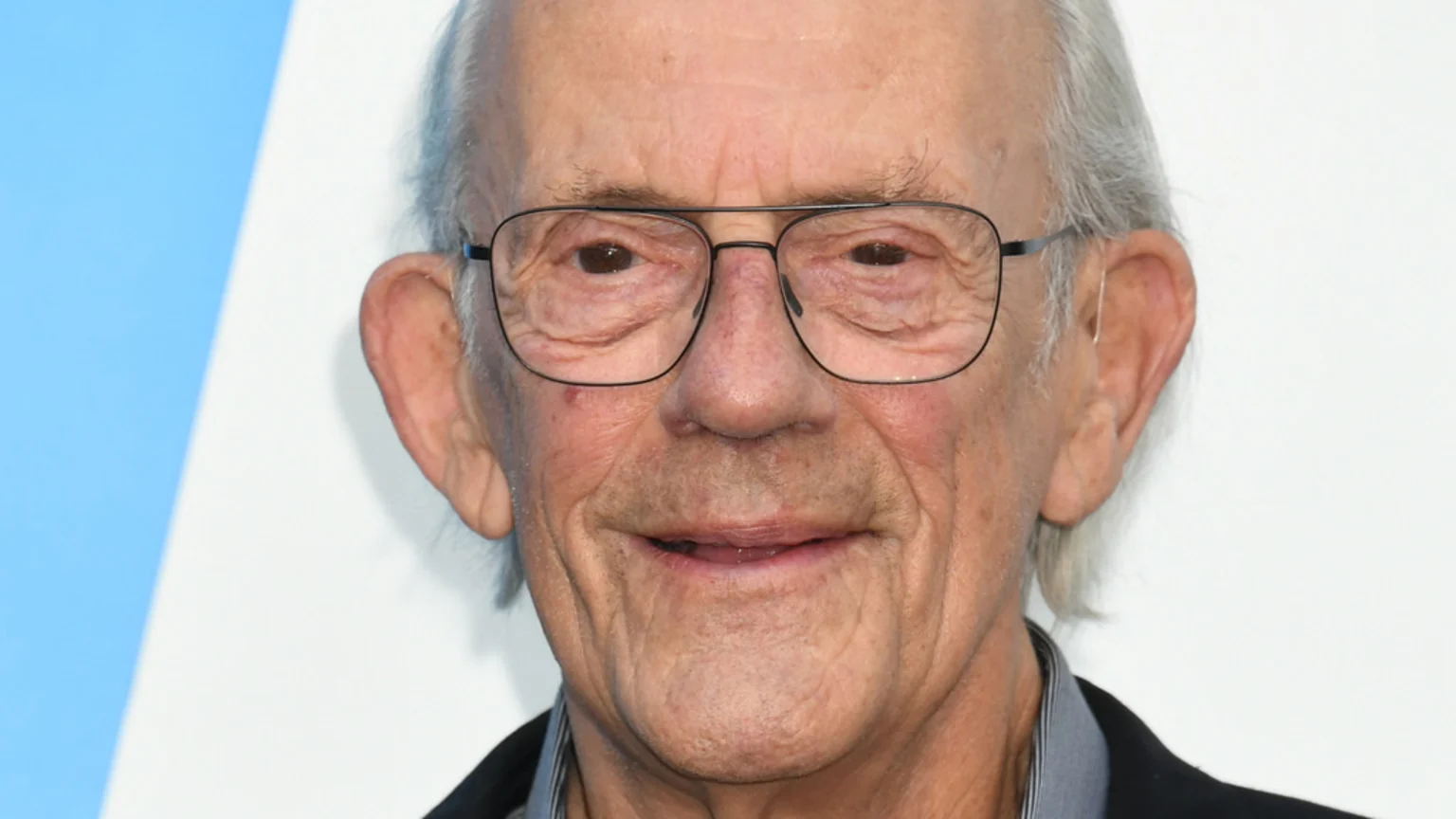 A close-up portrait of actor Christopher Lloyd with gray hair and glasses, smiling faintly and looking directly at the viewer against a blue and white background.