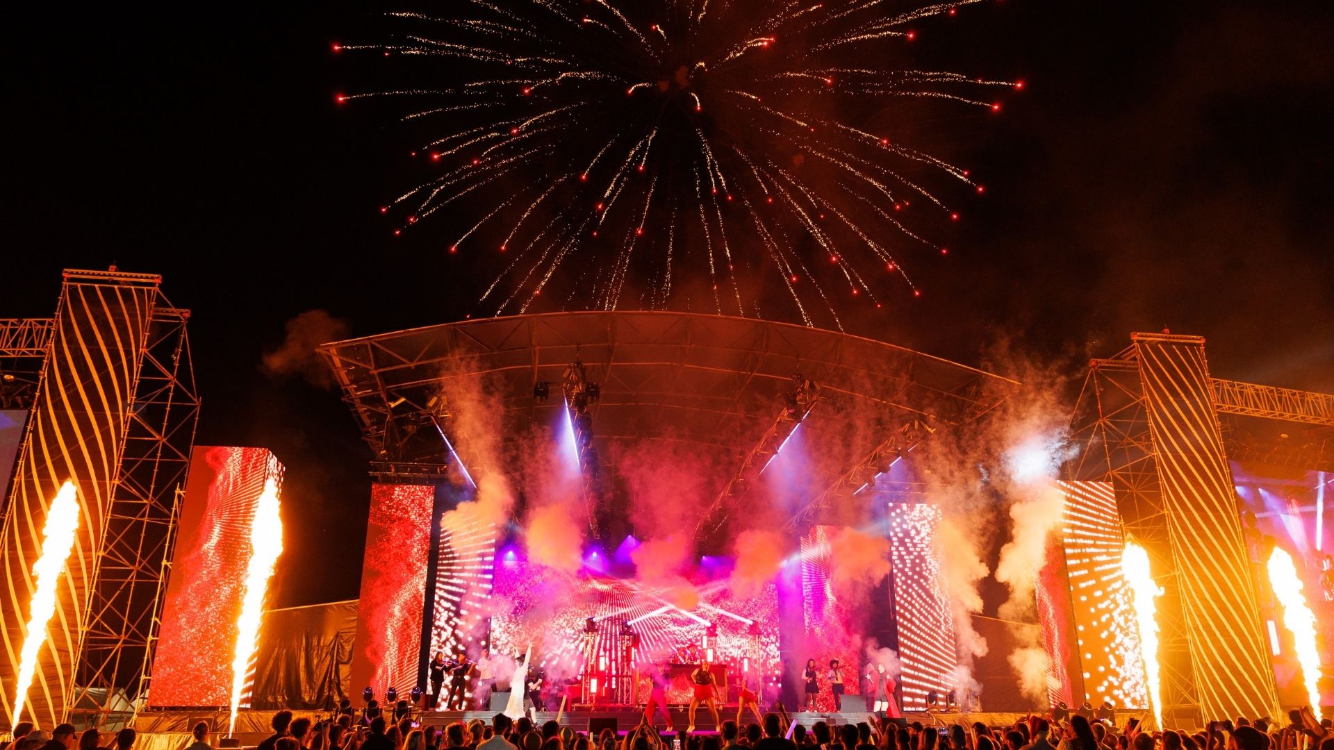 A festive outdoor concert at night, featuring a stage with dancers, colorful lights, flame jets, and fireworks exploding overhead, with a crowd watching.