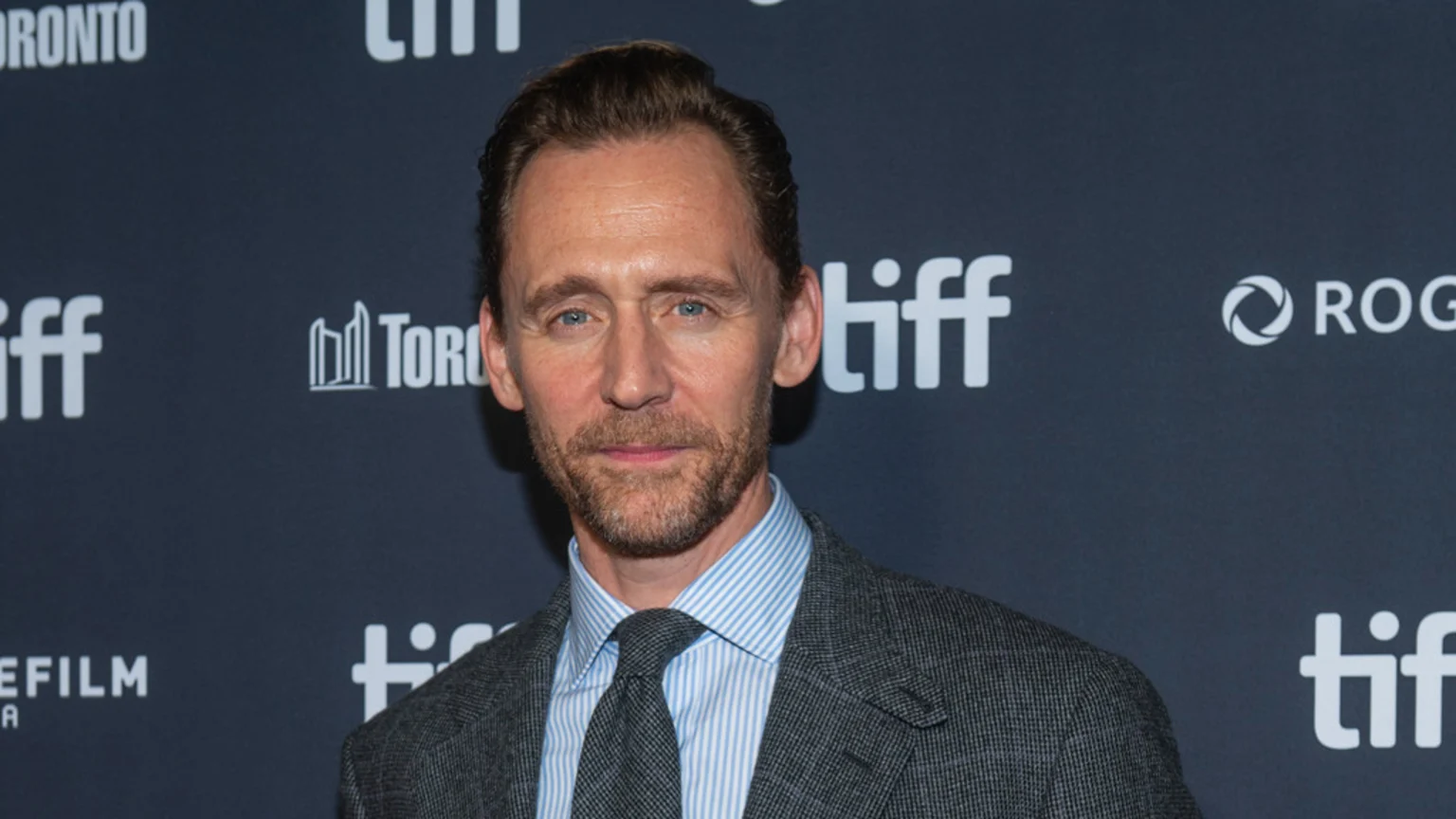 Actor Tom Hiddleston with light brown hair and a beard, wearing a striped shirt, tie, and grey suit jacket, poses in front of a dark background with white "TIFF" logos.