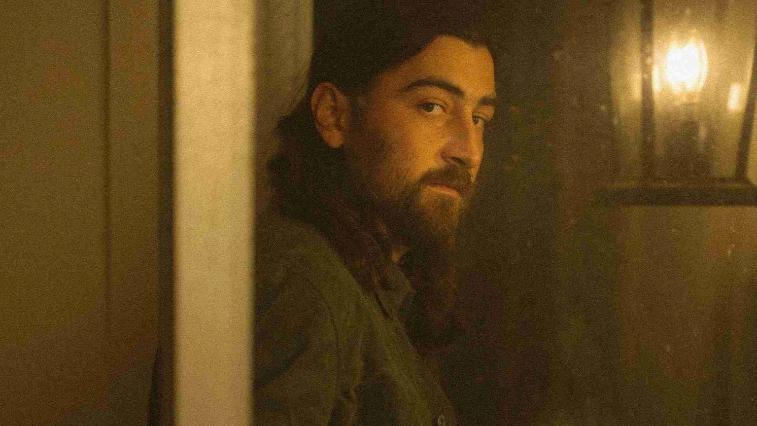 A bearded man with dark hair looks over his right shoulder towards the viewer, illuminated by warm light, with a bright light source in the blurry background.