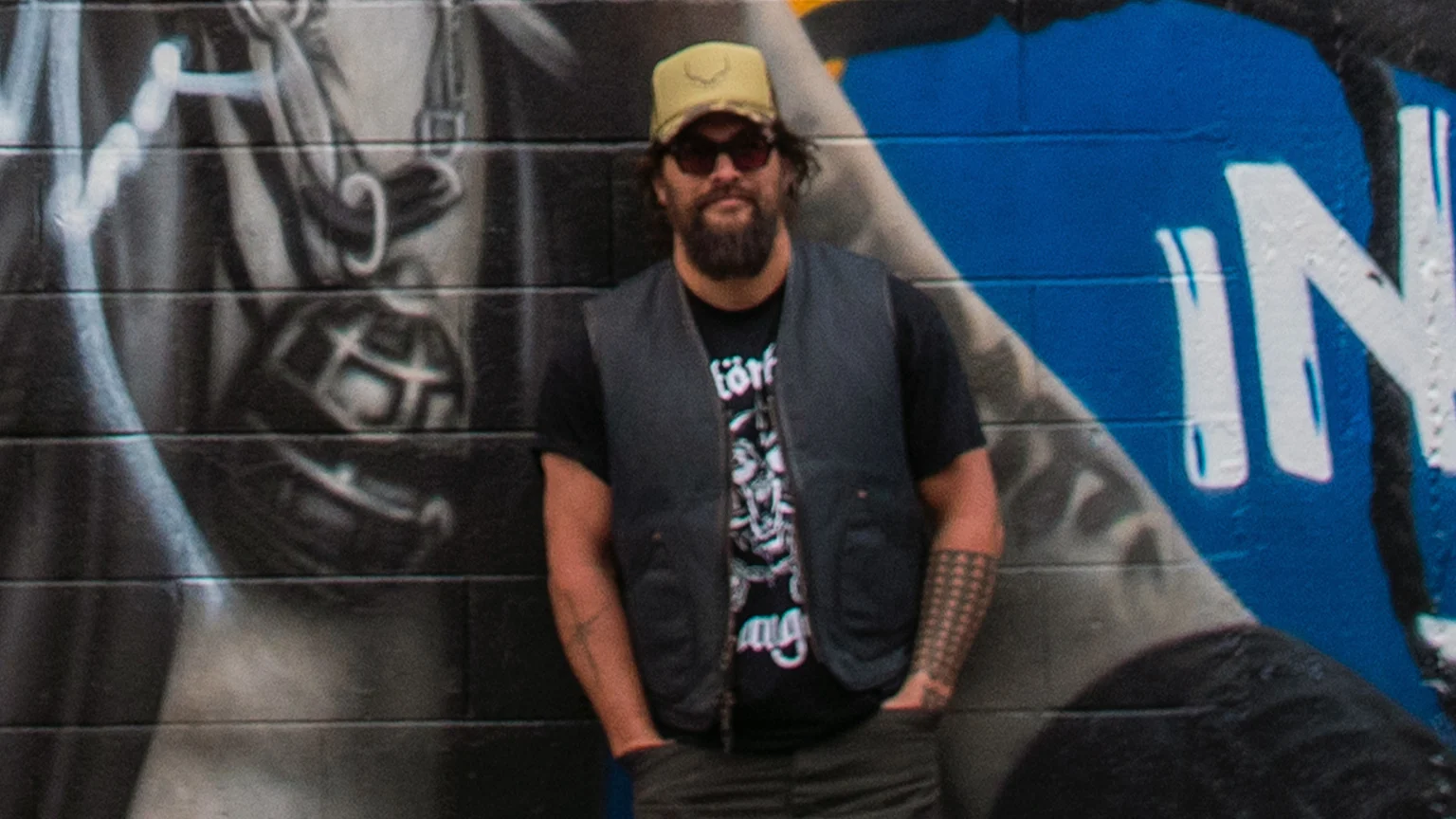 A bearded man in a cap, sunglasses, black t-shirt, and dark vest stands with hands in pockets before a street mural, featuring a black and white drawing on the left and a blue section with white letters on the right.