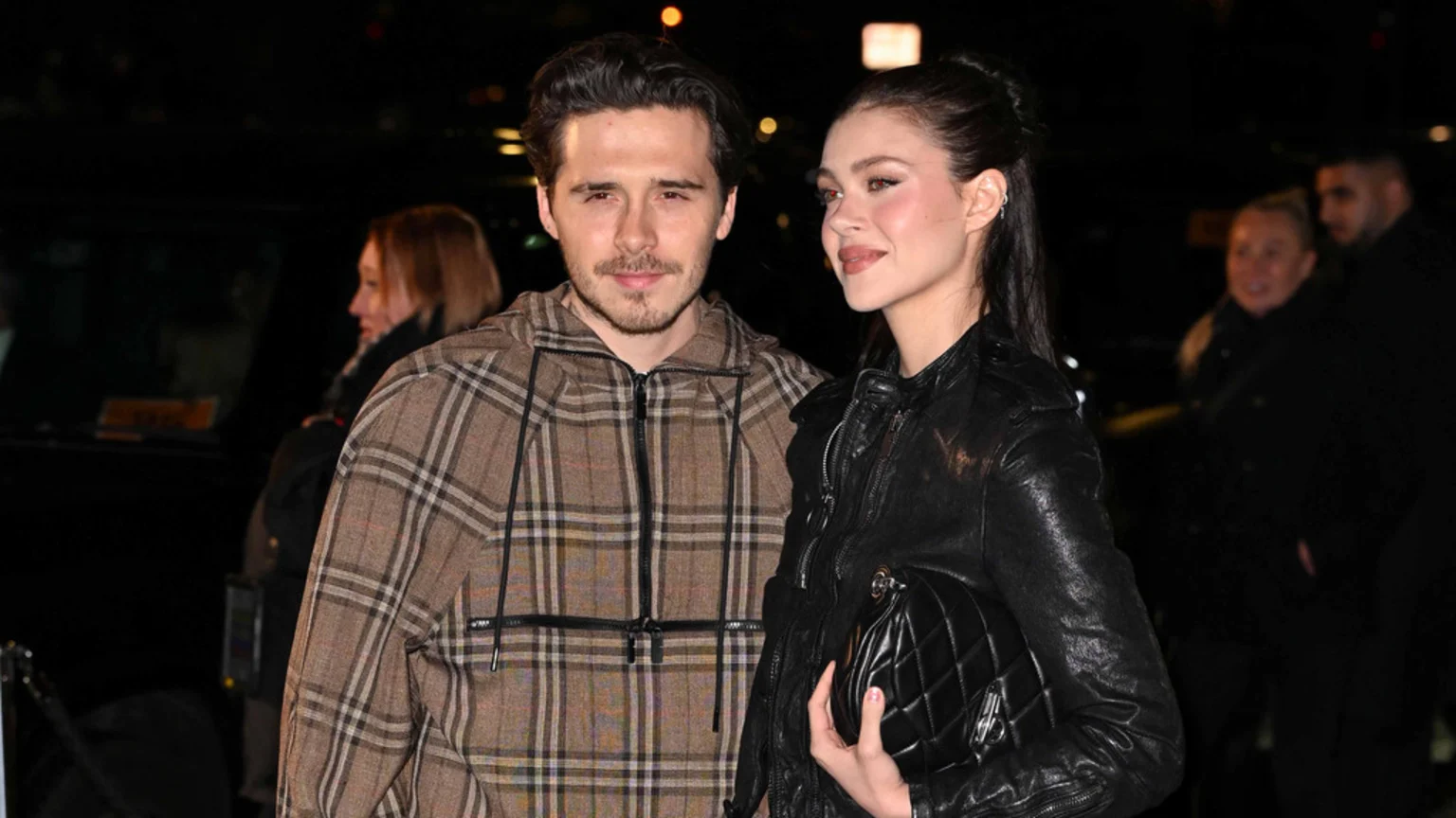 A man with a beard in a brown plaid jacket looks at the camera next to a woman with dark hair in a black leather jacket, holding a quilted bag, and looking slightly to the right with a subtle smile.