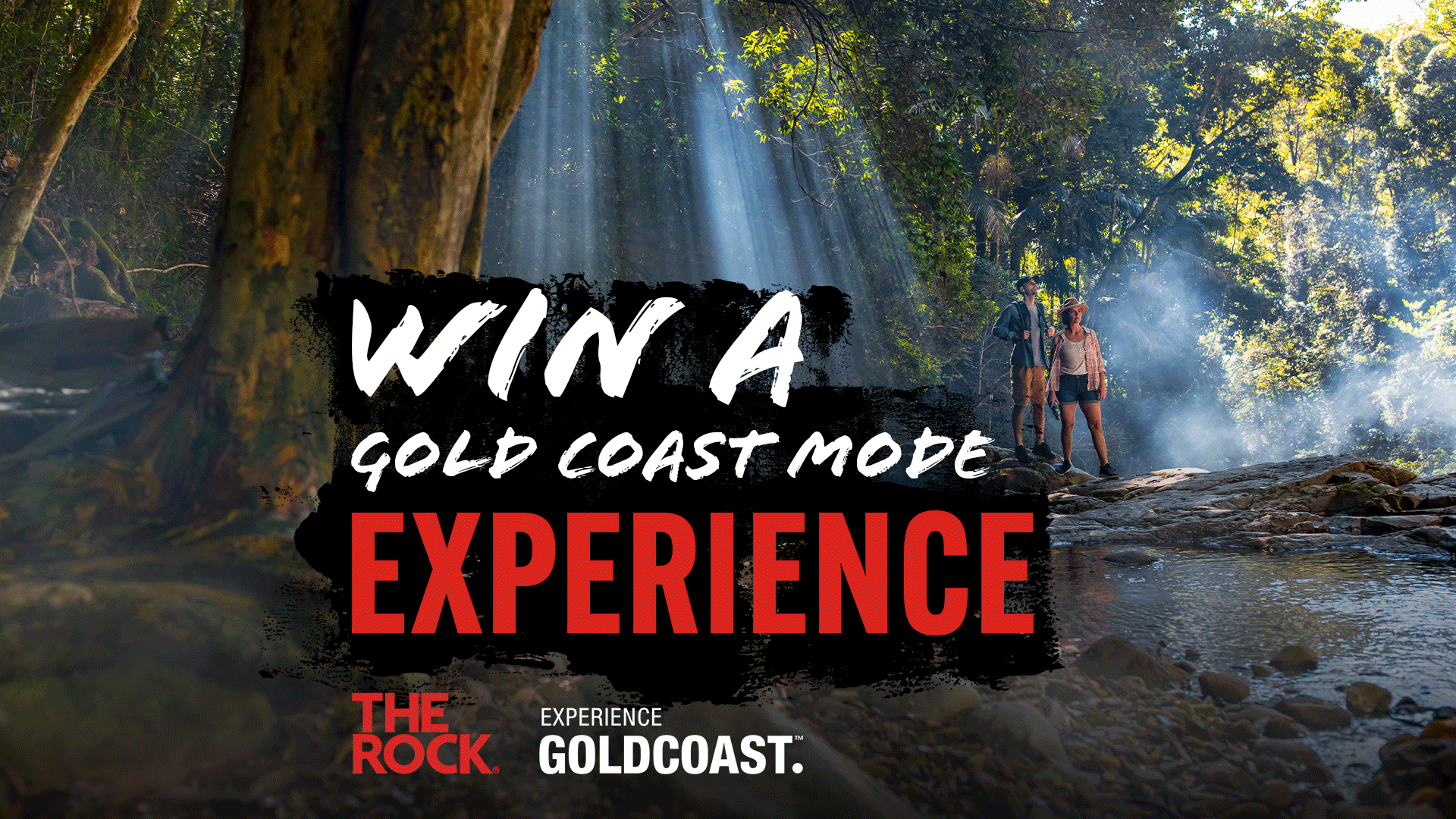 Two hikers stand on rocks next to a stream in a sun-drenched, misty forest, with text "WIN A GOLD COAST MODE EXPERIENCE" and "EXPERIENCE GOLDCOAST." logos.
