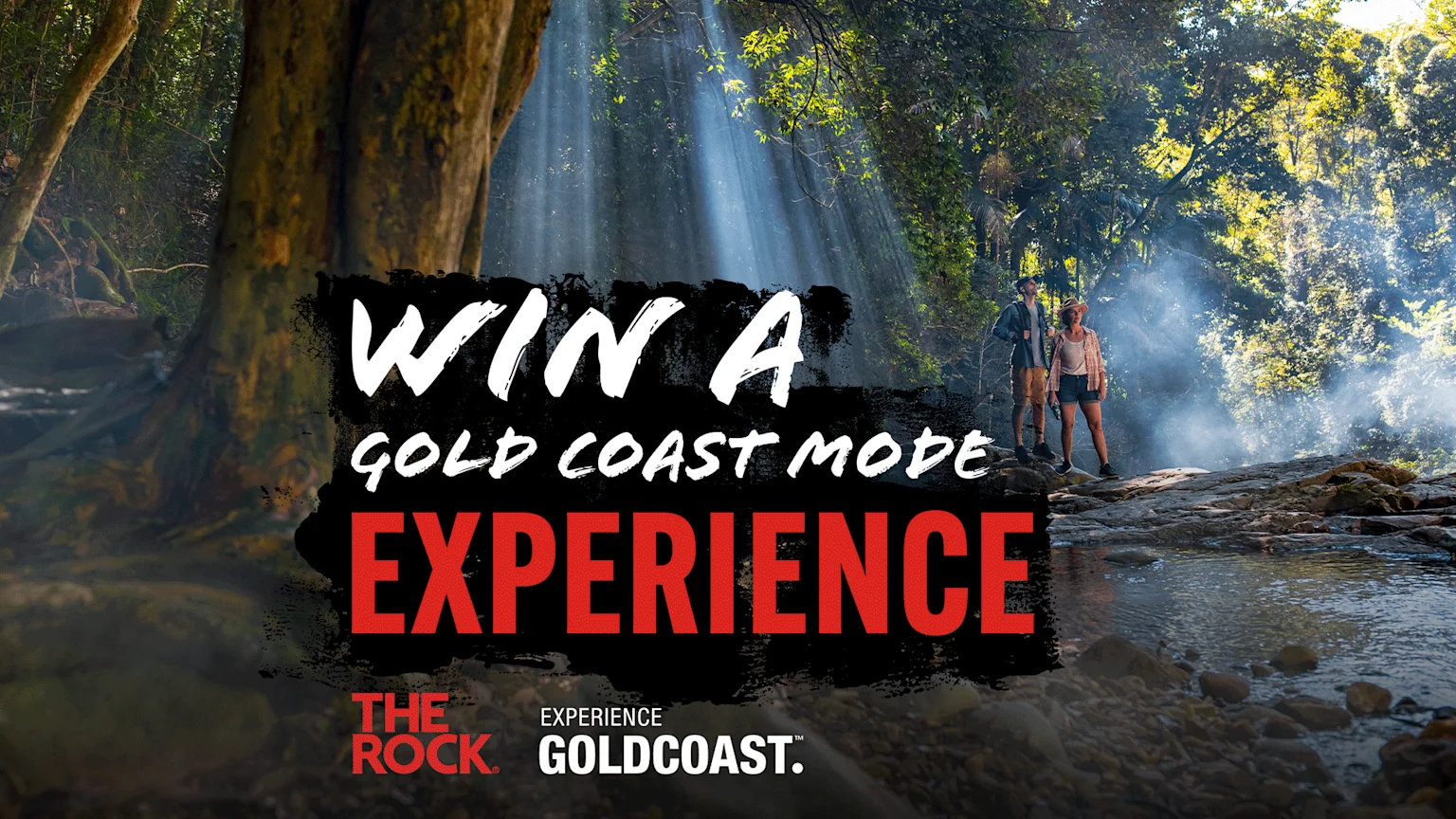 Two hikers stand on rocks next to a stream in a sun-drenched, misty forest, with text "WIN A GOLD COAST MODE EXPERIENCE" and "EXPERIENCE GOLDCOAST." logos.