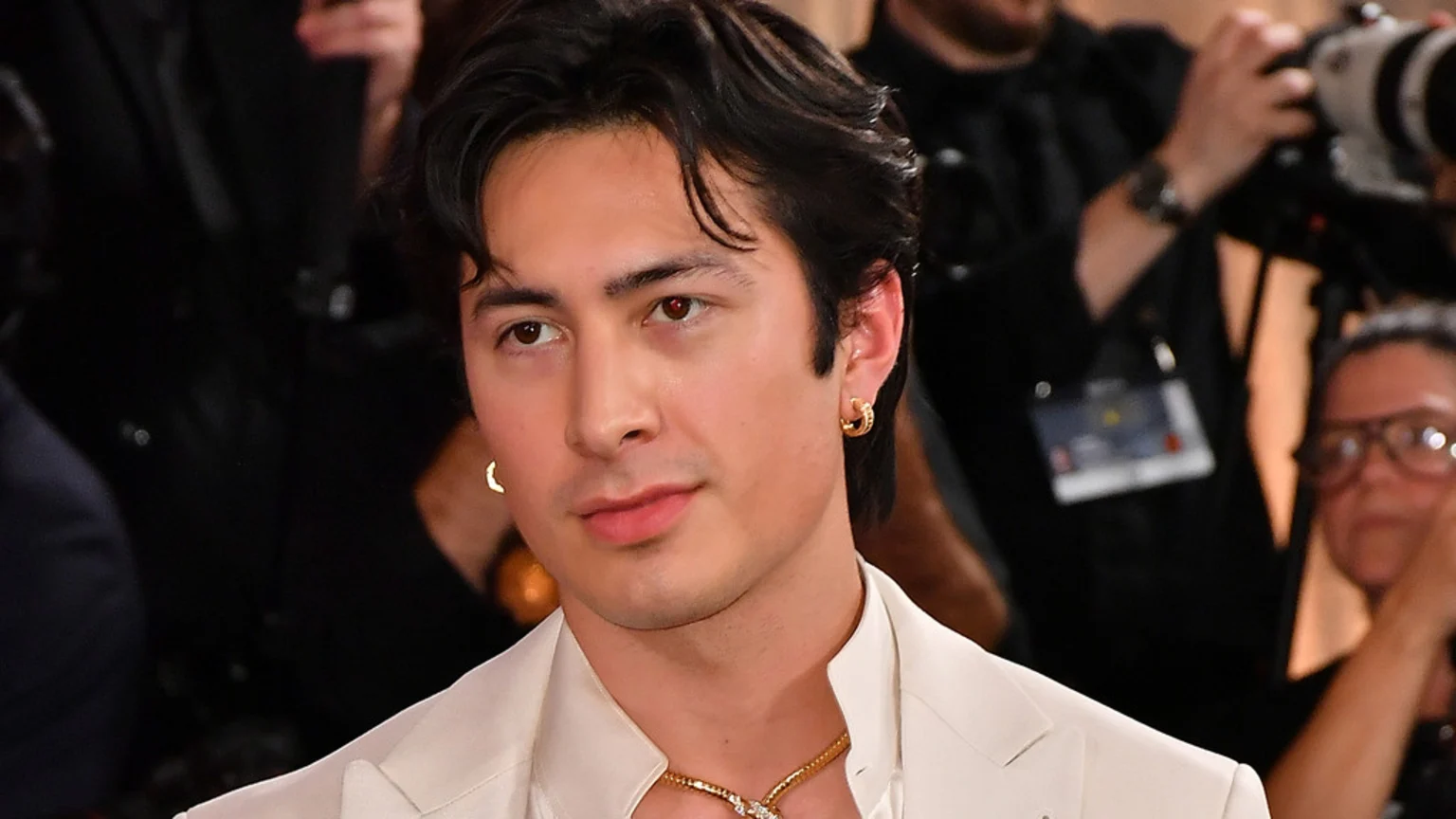 A man with dark wavy hair and light brown eyes wears a cream suit, gold hoop earrings, and a gold necklace, looking slightly left with a subtle smile.