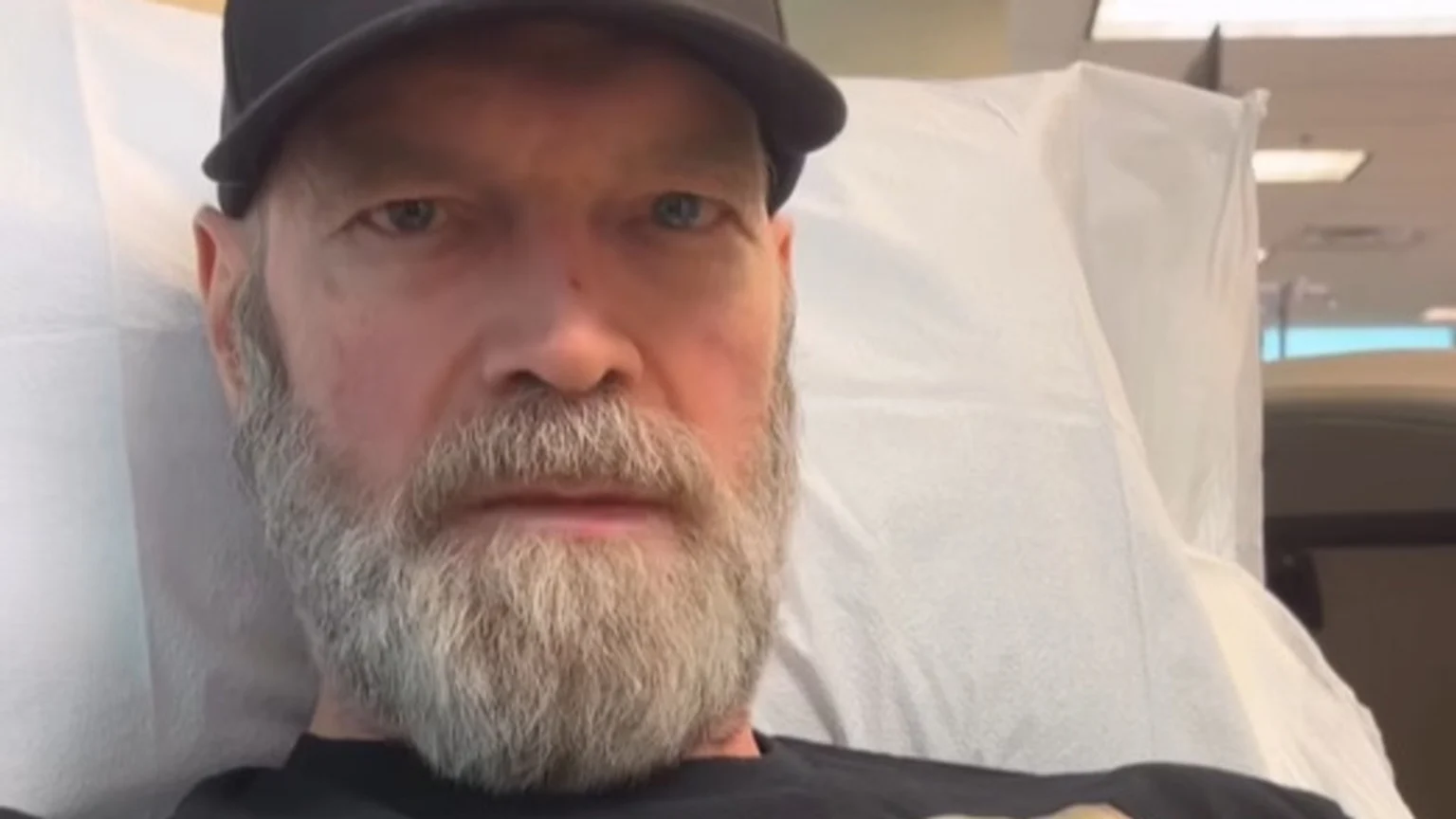A bearded man in a black cap lies on a white medical bed, looking at the camera.