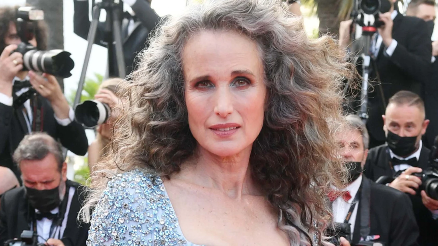 Andie MacDowell, with long grey curly hair and a sparkling light blue dress, smiles at the camera with photographers in the background at an event.
