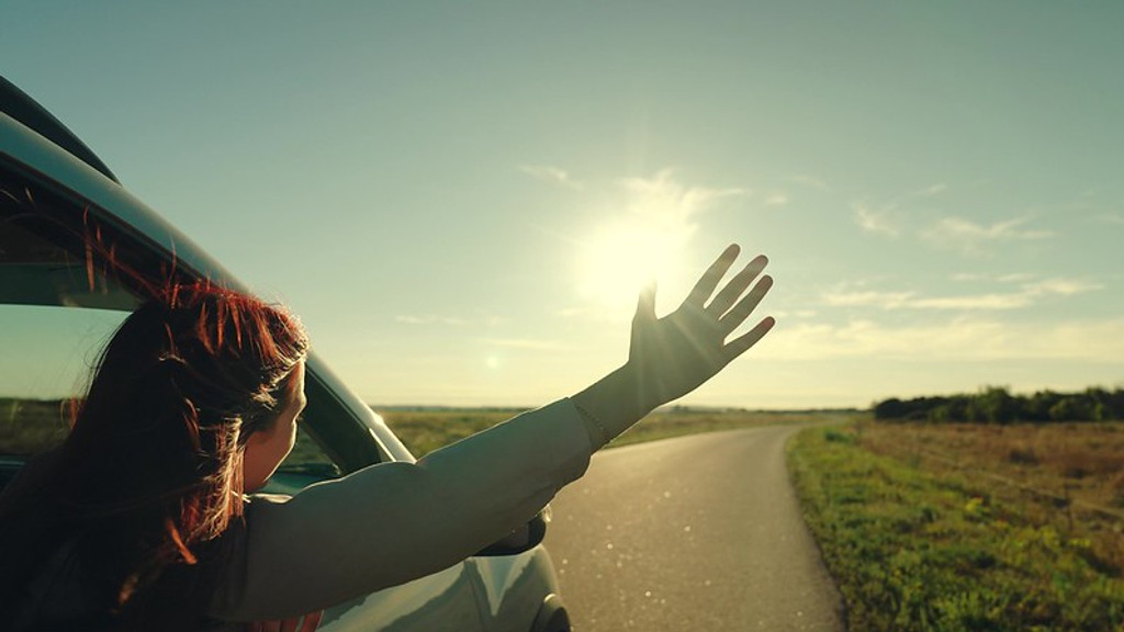 A person with reddish hair leans out a car window, extending a hand towards the bright sun in a clear sky above a rural road with green fields.