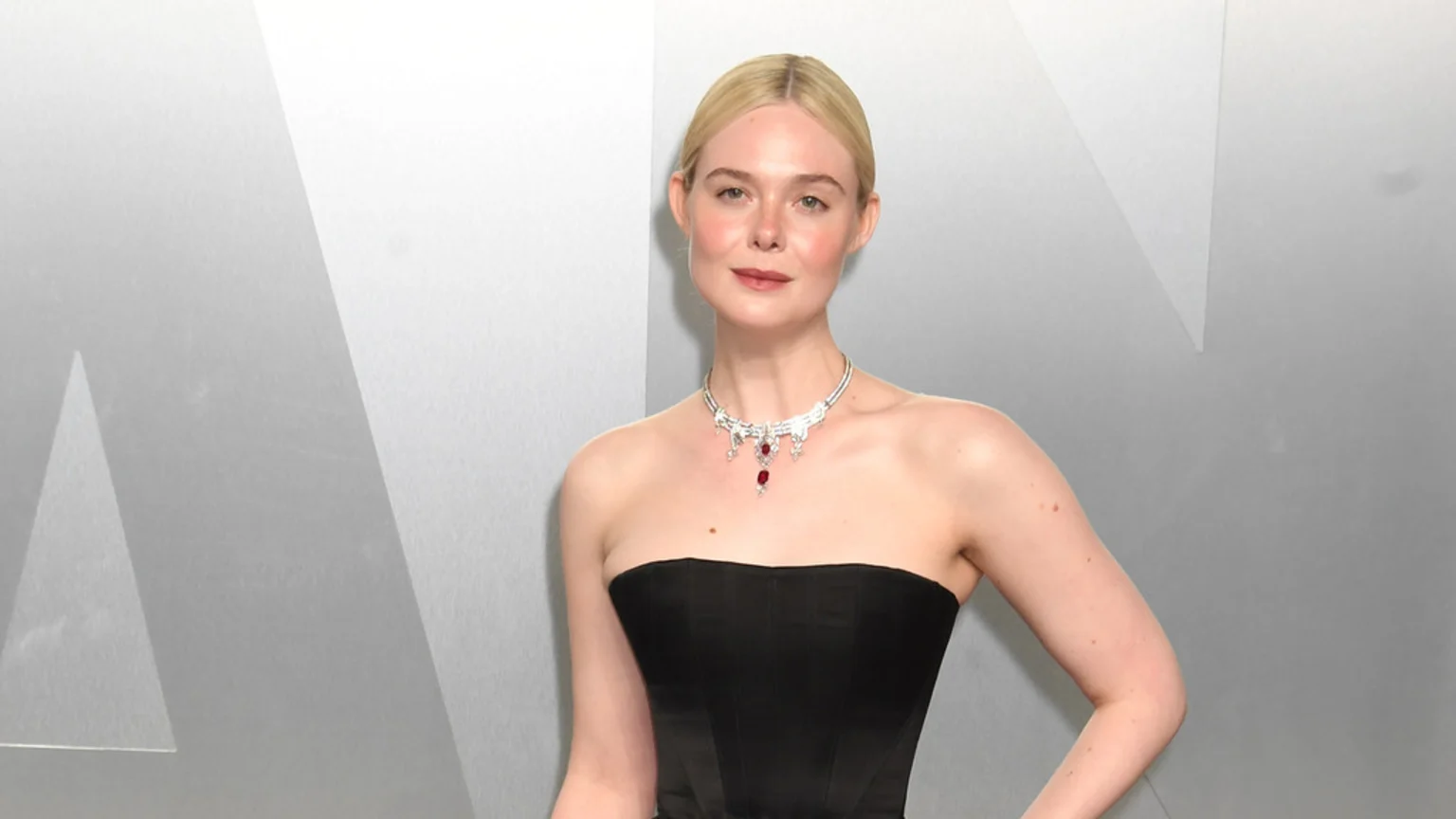 A woman with blonde hair in a sleek bun wears a black strapless dress and a silver necklace with red gems, posing against a light gray geometric background.