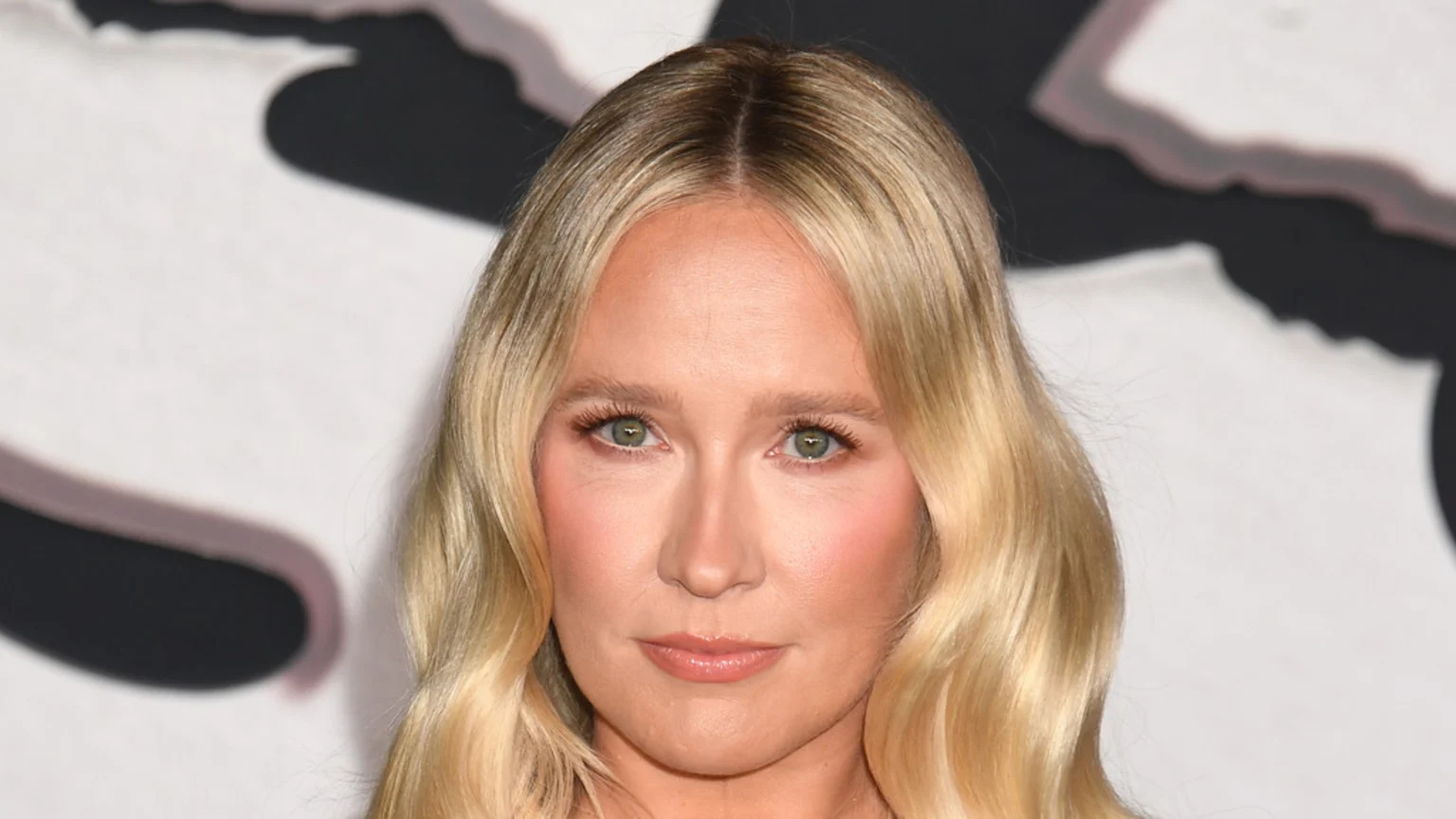 A close-up portrait of a woman with long, wavy blonde hair and green eyes, looking forward with a neutral expression. She wears natural-looking makeup, against a blurry white background with dark shapes.