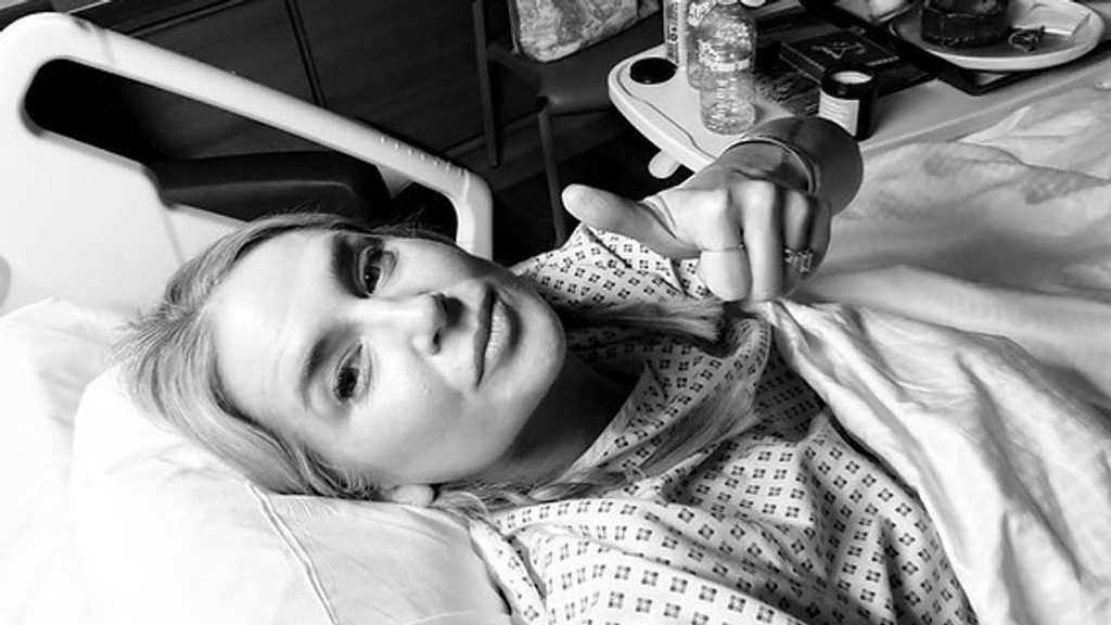 A woman in a hospital gown lies in bed, looking at the camera and pointing her right hand, wearing rings, with a bedside table in the background, in a black and white shot.