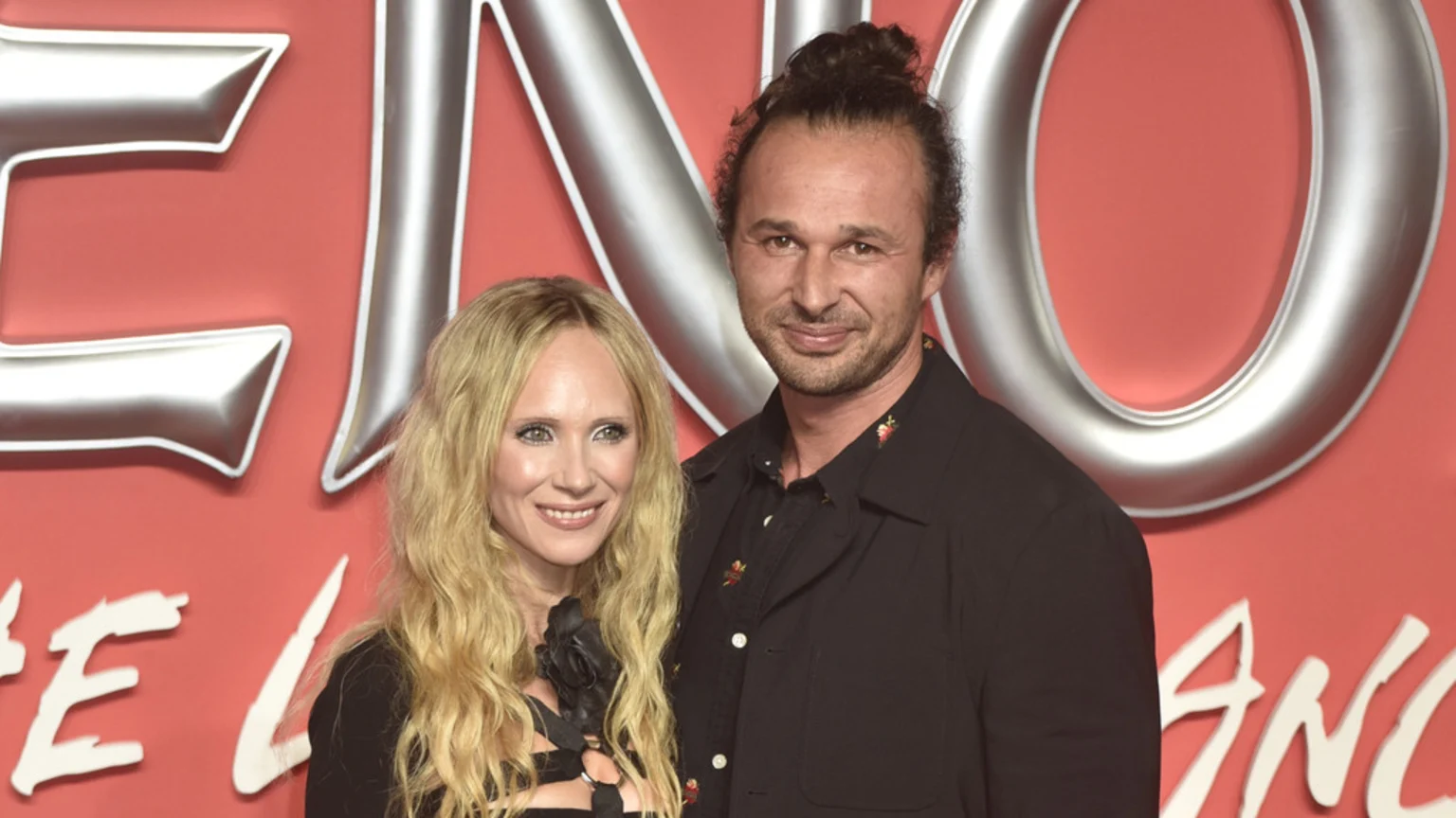 A smiling woman with long blonde hair and a man with a bun stand together in front of a red background with large silver letters.