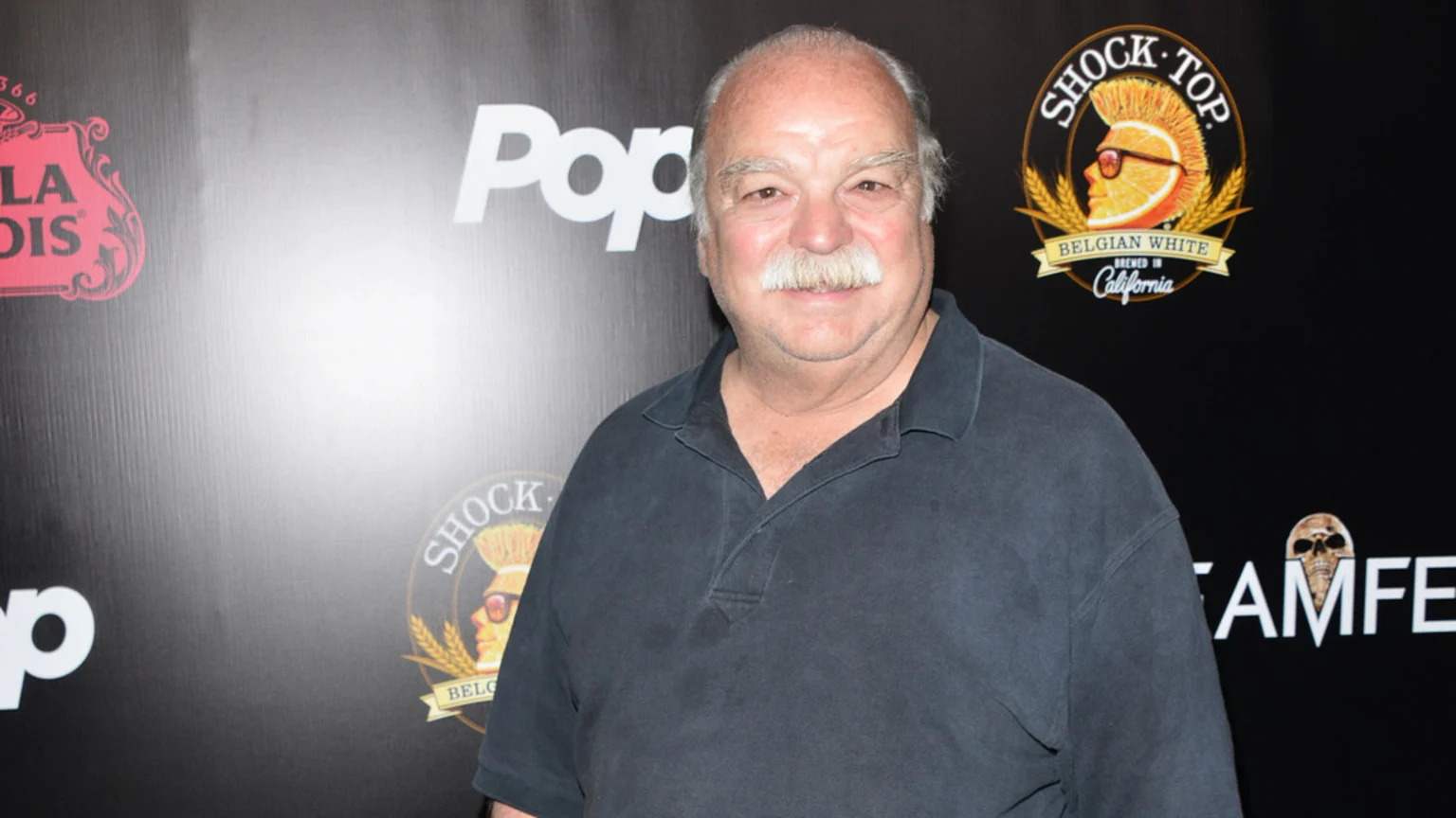 A man with white hair and a mustache, wearing a dark polo shirt, smiles at the camera in front of a black background featuring logos for Shock Top and Stella Artois.