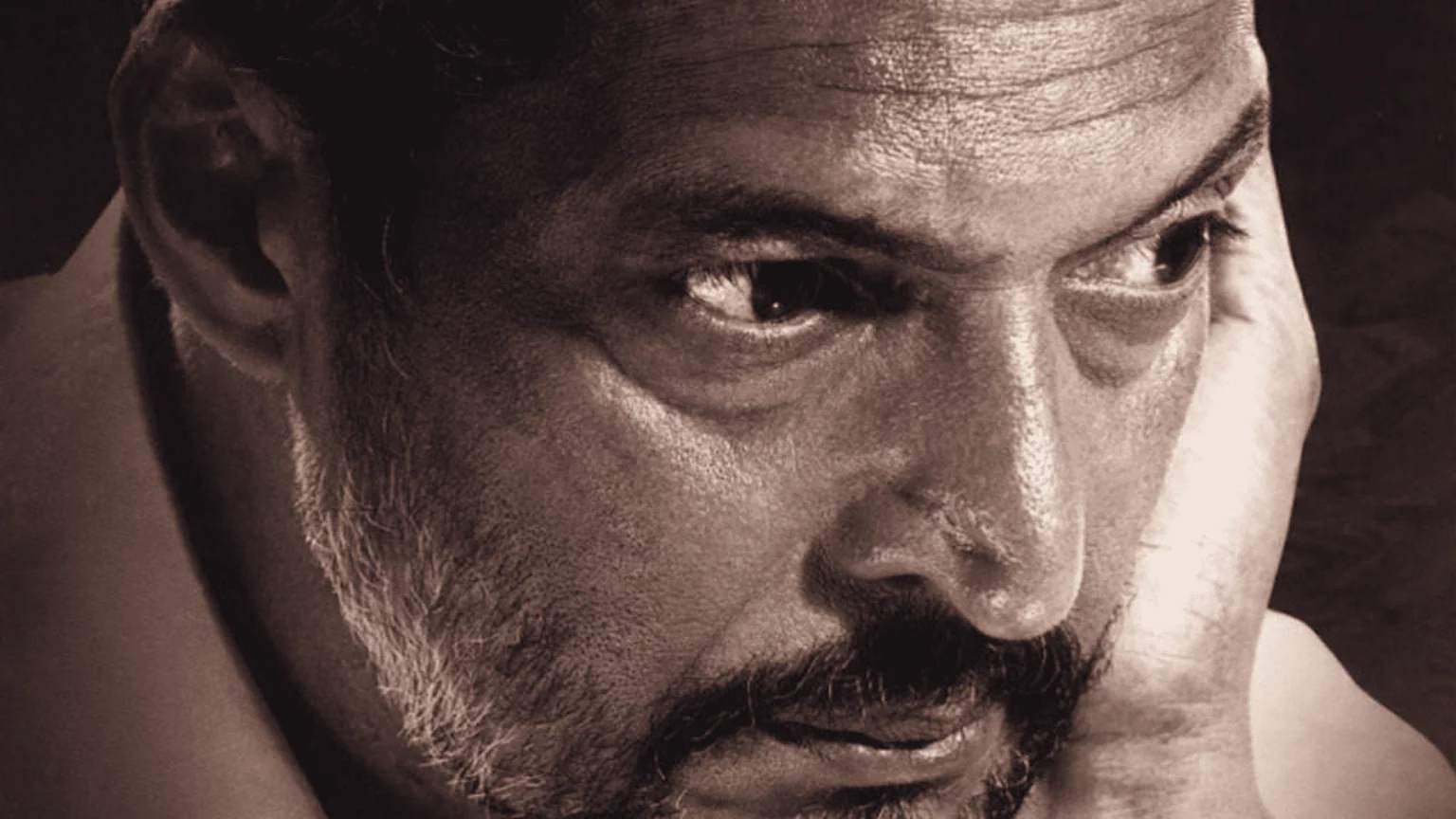A sepia-toned close-up portrait of a man with a beard and mustache, looking intensely to the right, with his hand resting on his face, suggesting deep thought.