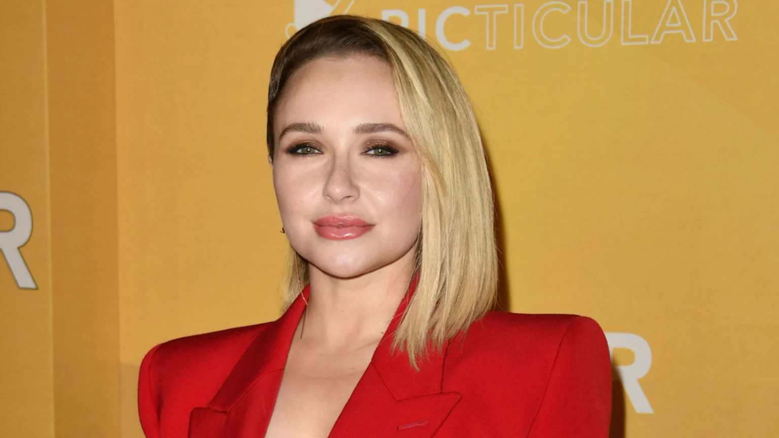 A woman with blonde hair and green eyes, wearing makeup and a red blazer, stands against a yellow background with partial white text visible.
