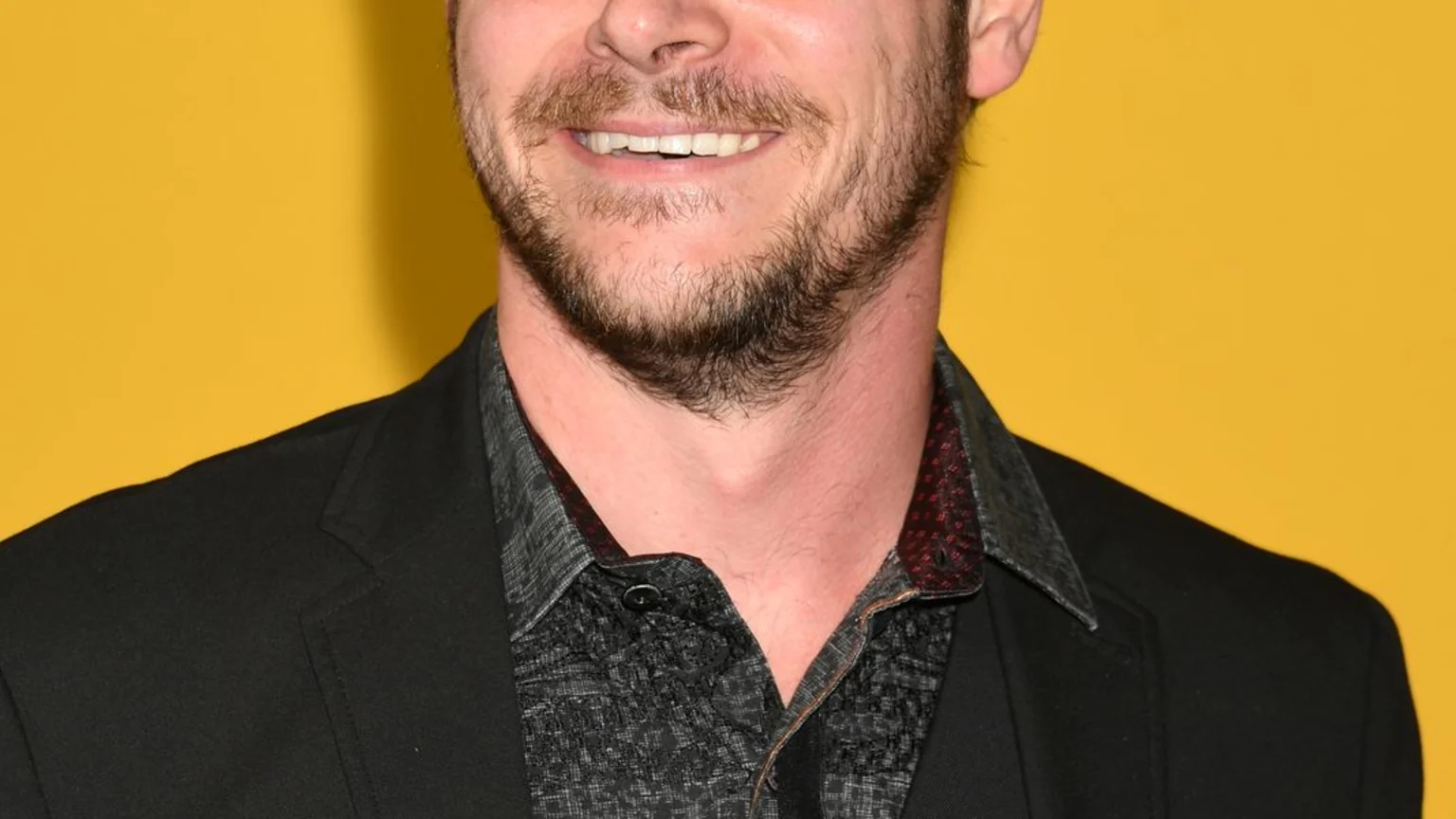 A smiling man with dark, messy hair and a beard wears a dark patterned shirt, vest, and black suit jacket against a bright yellow background with blurry yellow letters.