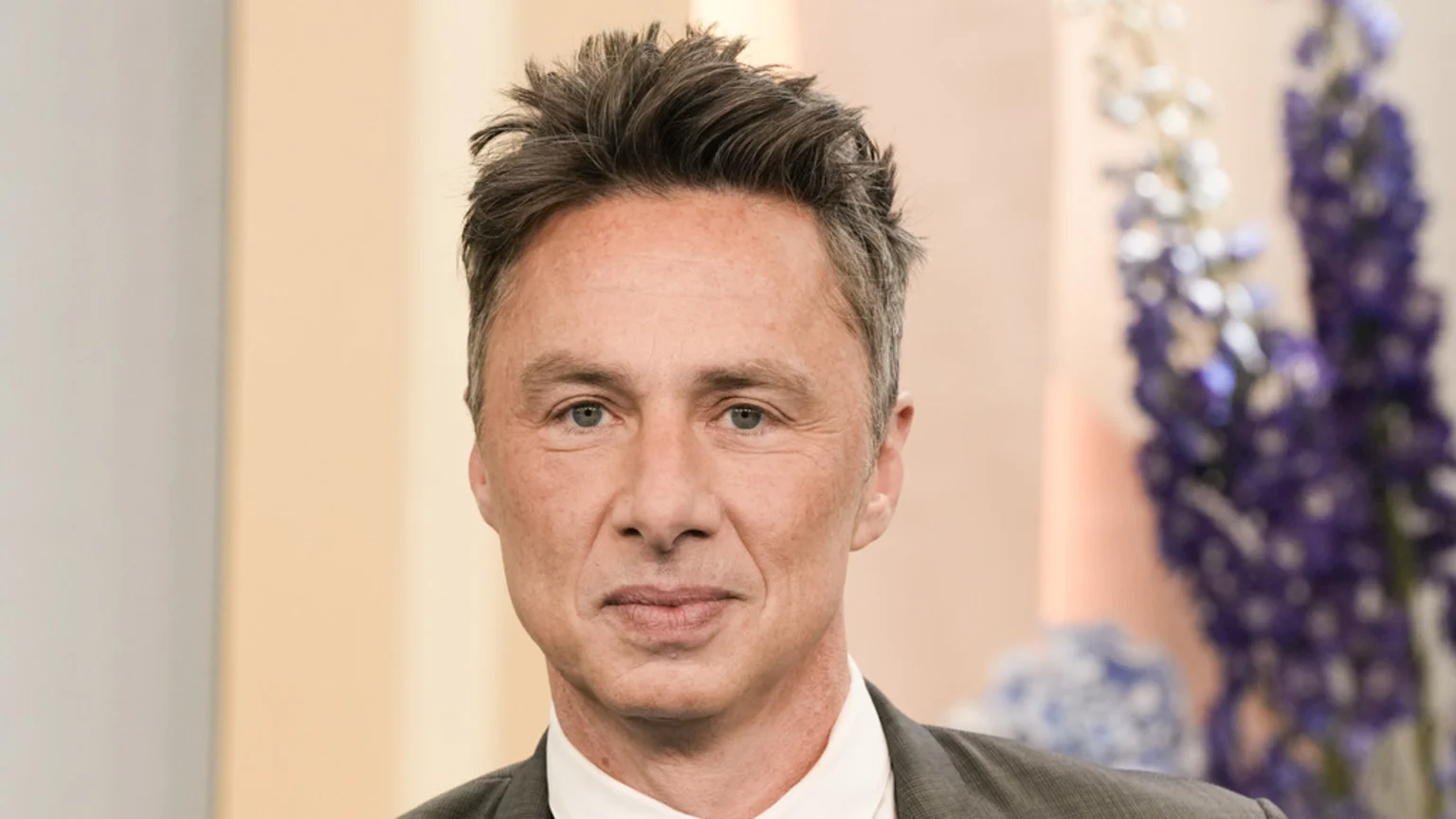 A man with short, spiky grey hair and light eyes looks directly at the camera, wearing a dark suit jacket and white shirt against a blurred light background with hints of purple flowers.
