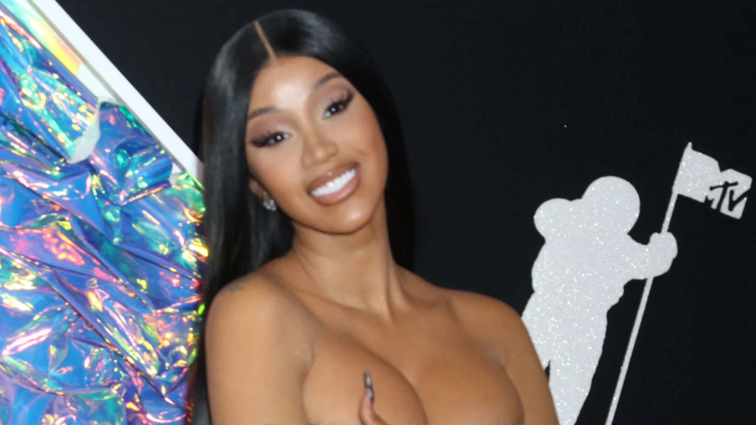 Cardi B smiles, showing her shoulders and cleavage, with long dark hair, against a bright blue iridescent fabric and a white glitter MTV astronaut silhouette.