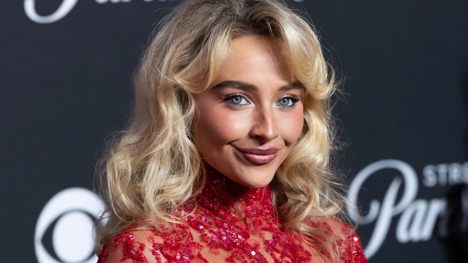 Sabrina Carpenter smiles subtly at the camera, wearing a high-necked, red sequined top and sporting blonde wavy hair, with blue eyes and a dark background.