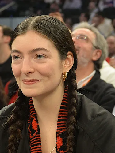 Singer Lorde smiles gently, wearing her hair in two braids and gold hoop earrings, while seated among other spectators who are looking up at an event.