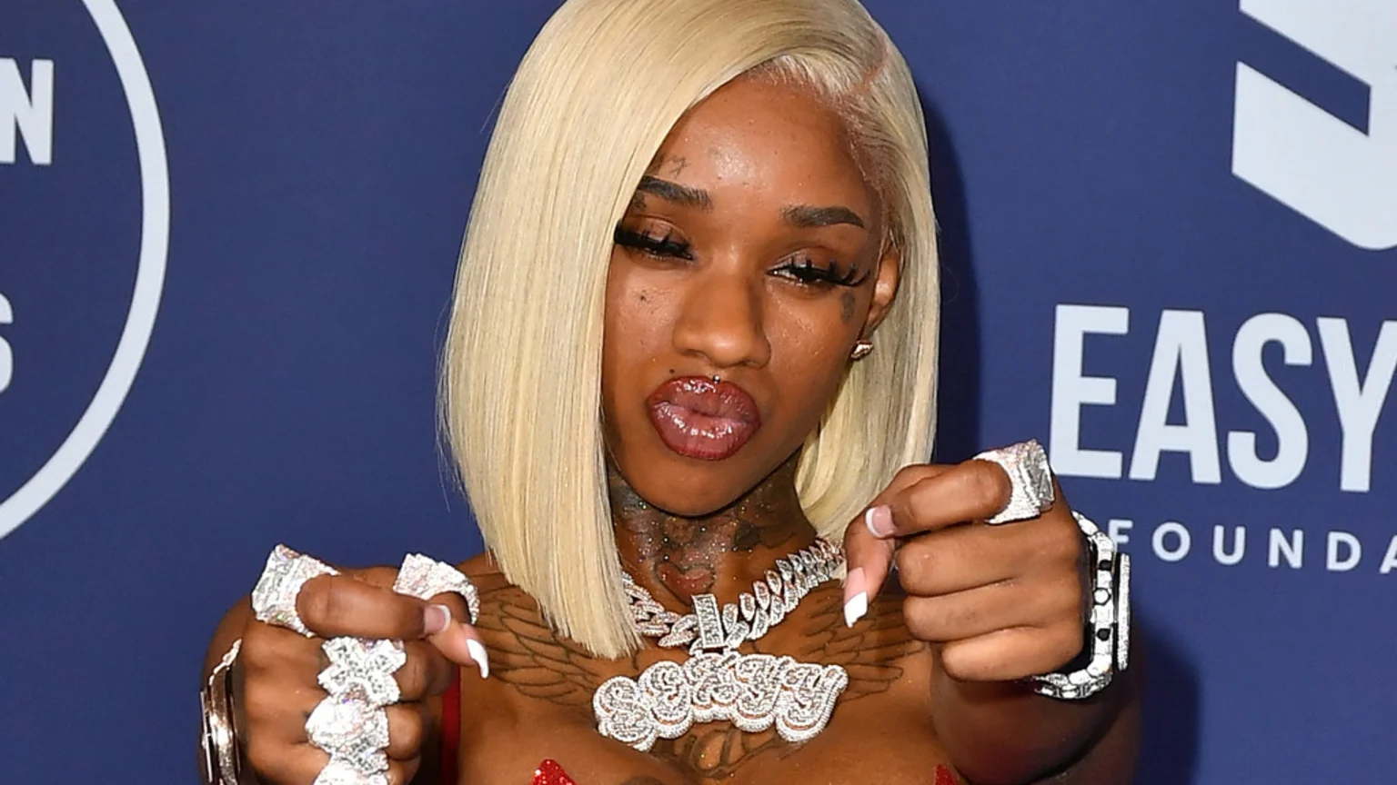 A woman with platinum blonde bob hair, facial and neck tattoos, and a lip piercing, wearing a diamond-encrusted custom nameplate necklace and multiple diamond rings, looks directly at the camera while presenting her hands forward against a blue background with the "EASY FOUNDATION" logo.