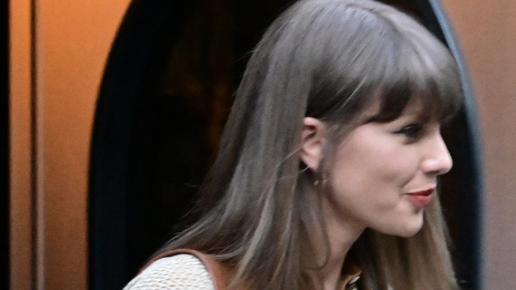 Close-up side profile of Taylor Swift with long brown hair, bangs, red lipstick, and eyeliner, looking right against a blurred warm-toned background.
