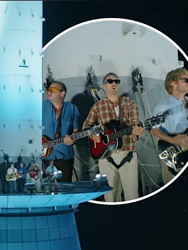 A group of six adult male band members perform on a tall illuminated tower platform at night. Three members in the foreground play guitars, wear sunglasses and safety harnesses, with city lights in the background.