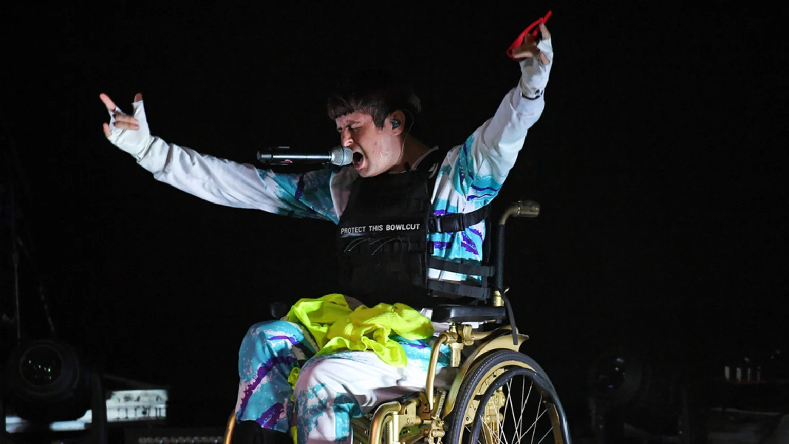 A person with a bowl cut, wearing a colorful patterned outfit and a vest that says "PROTECT THIS BOWLCUT," sings into a microphone while seated in a golden wheelchair on a dark stage.