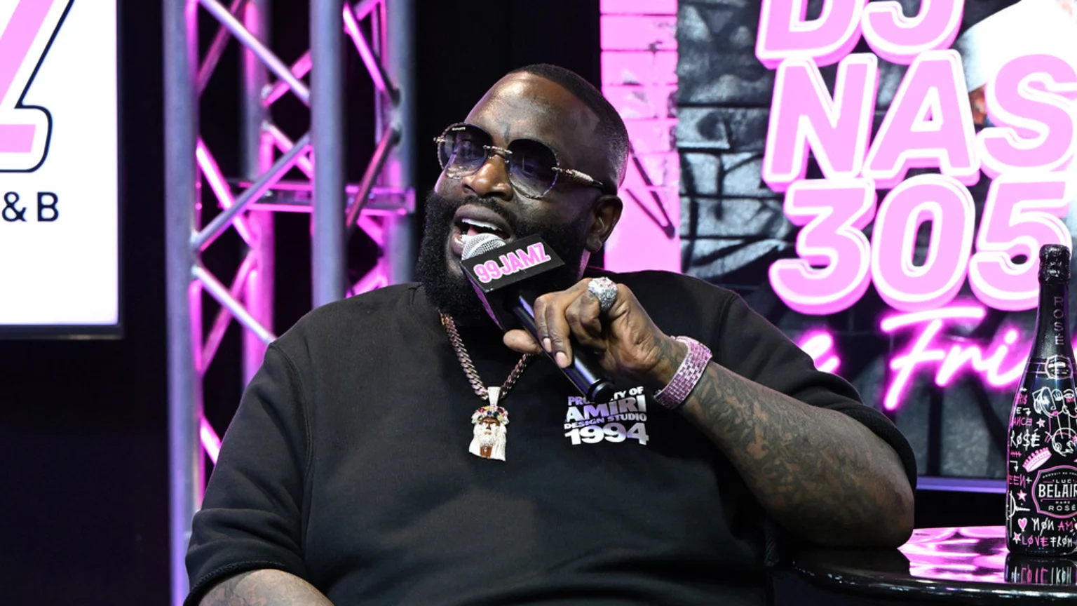 Rapper Rick Ross, wearing sunglasses, a black shirt, and jewelry, speaks into a "99 JAMZ" microphone while sitting indoors near pink neon signs and a "BELAIR ROSE" bottle.
