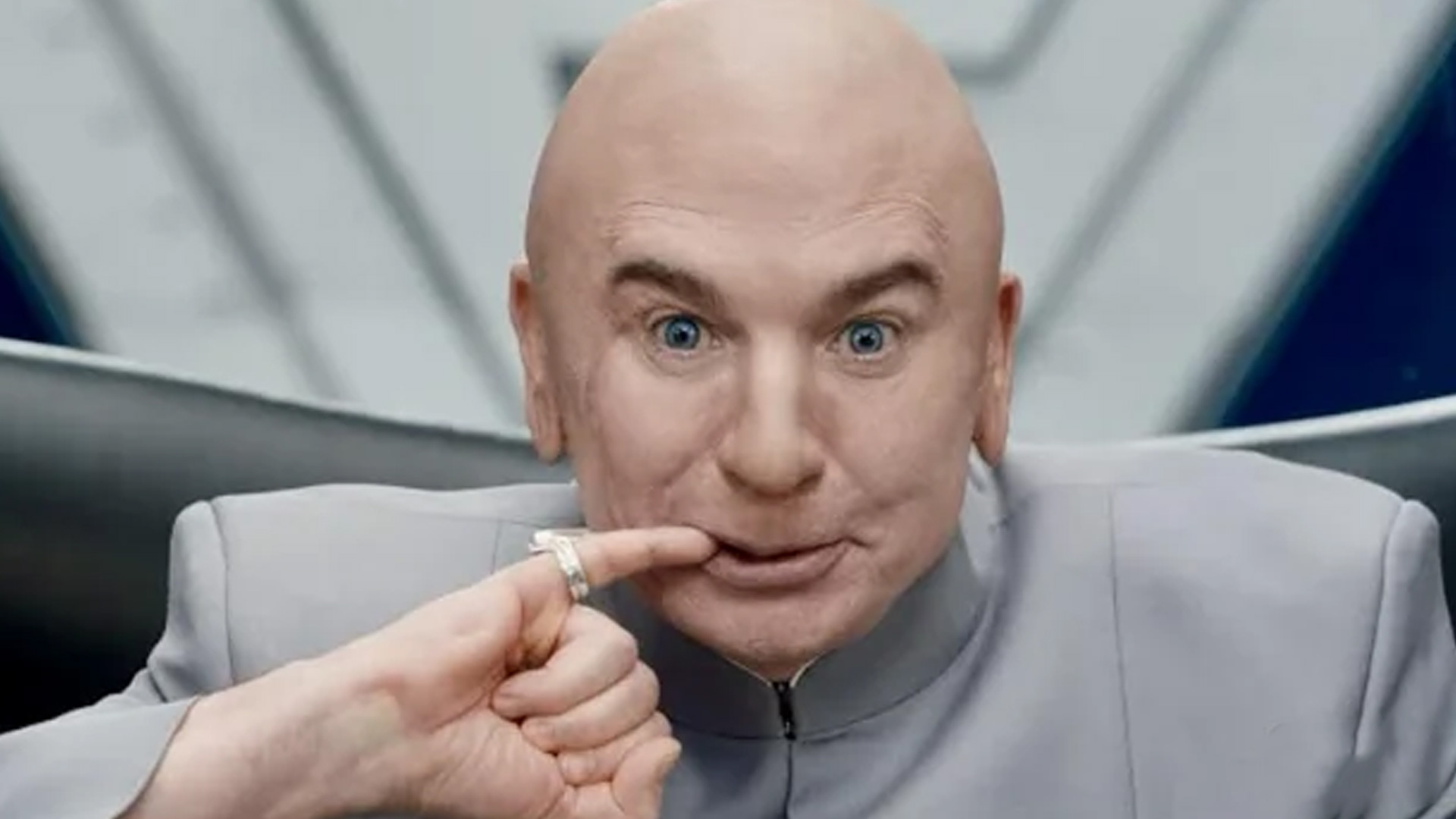Actor Mike Myers, with short platinum blonde hair, wears a dark suit and shirt, smiling subtly against a dark background with gold circular details.