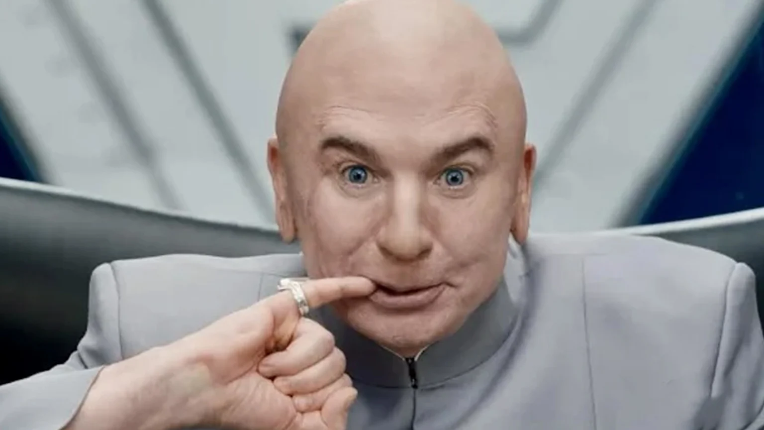 Actor Mike Myers, with short platinum blonde hair, wears a dark suit and shirt, smiling subtly against a dark background with gold circular details.