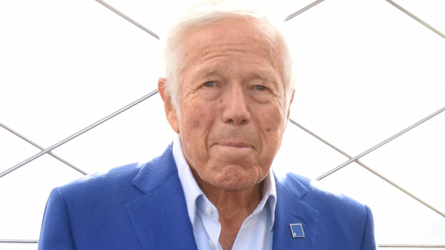 A close-up portrait of Robert Kraft, an older man with white hair, wearing a blue suit jacket and light blue shirt, looking directly at the camera, with a bright white background and diagonal metal bars.