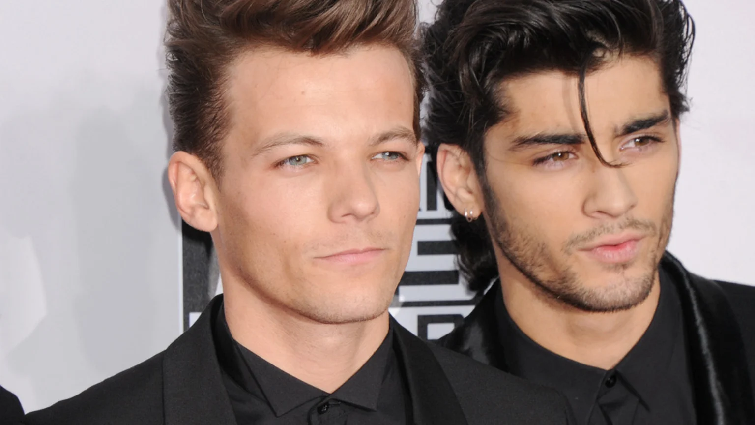 Close-up of Louis Tomlinson on the left with light brown hair and blue eyes, and Zayn Malik on the right with dark hair, brown eyes, and a beard, both wearing black.