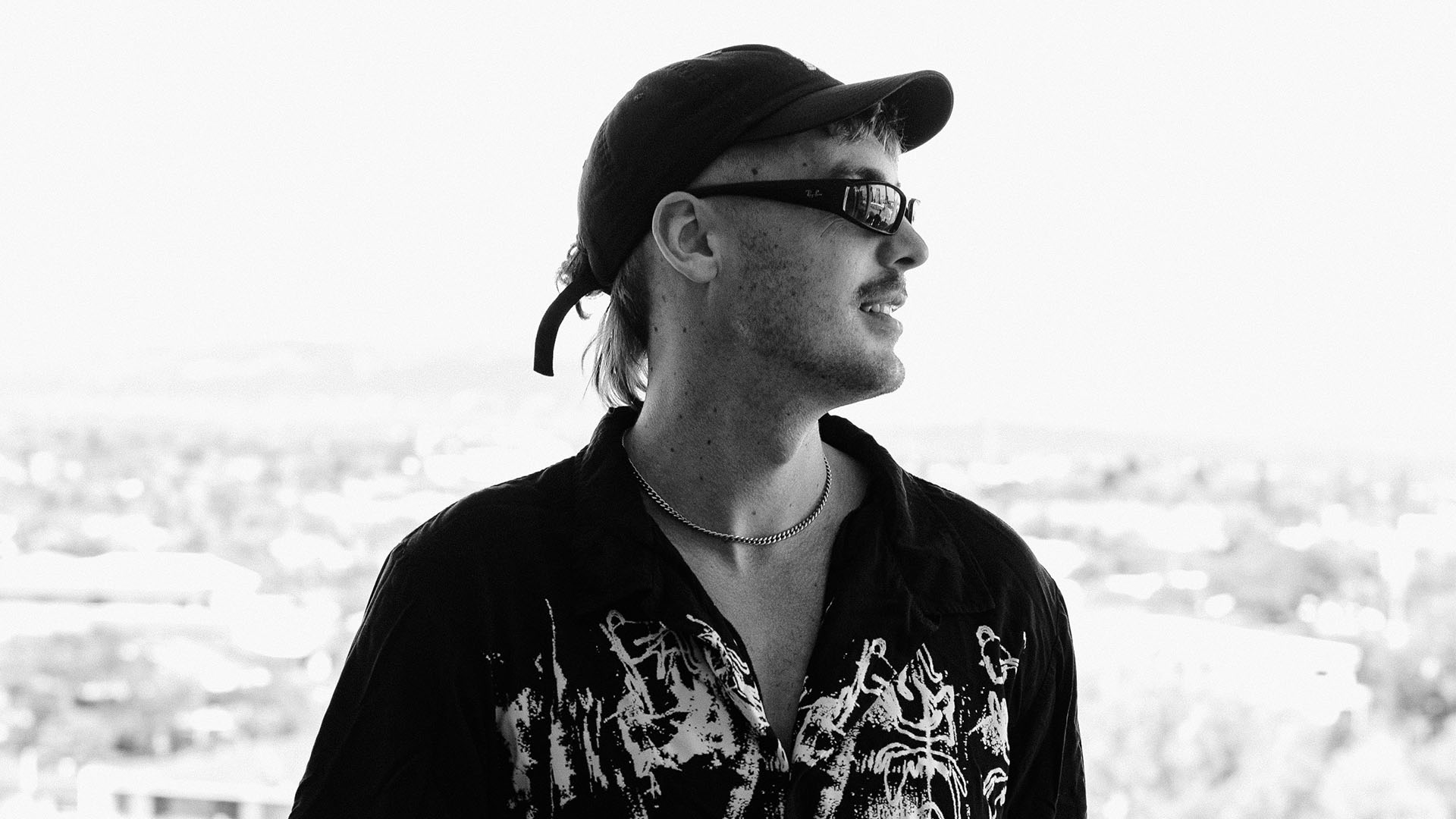 A black and white photo of a man with a mullet, wearing a backward cap, small sunglasses, a chain, and a patterned shirt, looking right with a faint smile against a bright, blurred city backdrop.