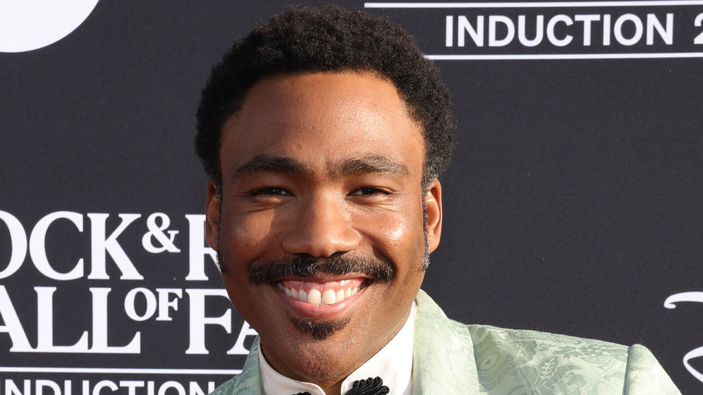 Donald Glover smiles broadly, showing his teeth, wearing a light green patterned suit jacket and bow tie, with a "Rock & Roll Hall of Fame" sign in the background.