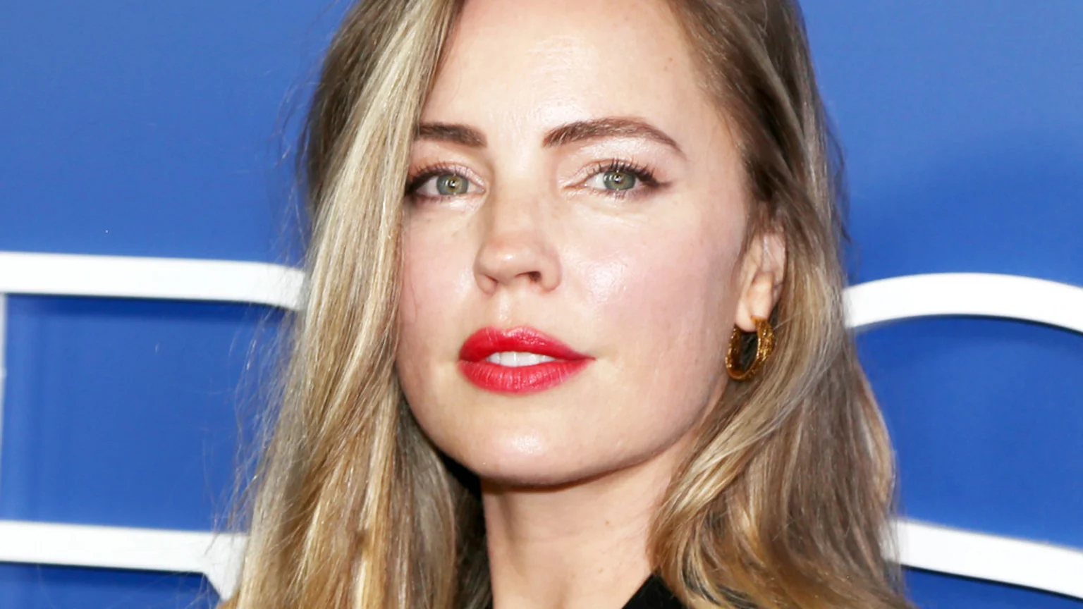 Close-up of a woman with blonde hair, green eyes, and red lipstick, wearing a gold hoop earring against a blue background with white lines.